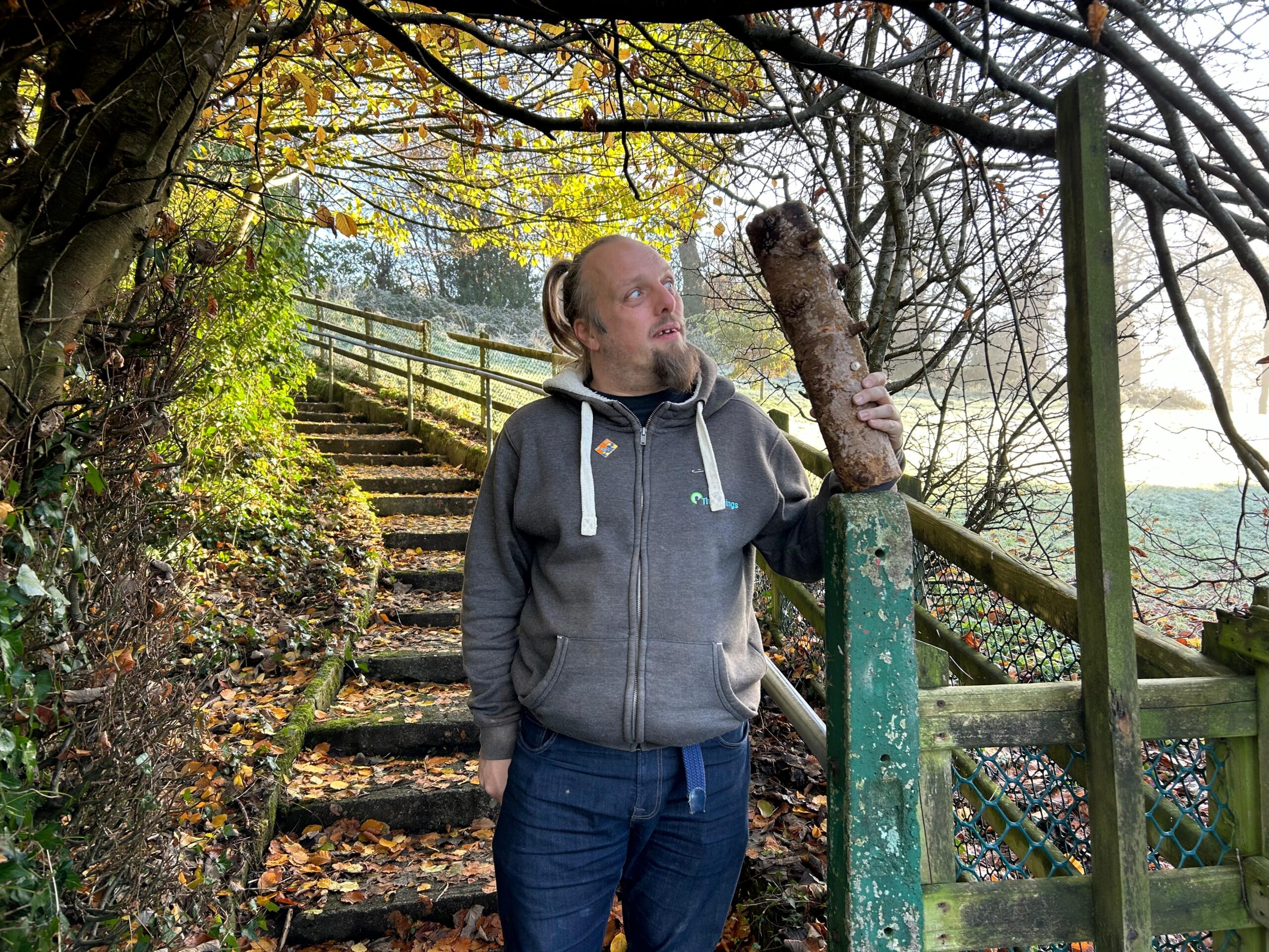 Full image of Dan stands by some steps in a park, holding a strange-looking log.