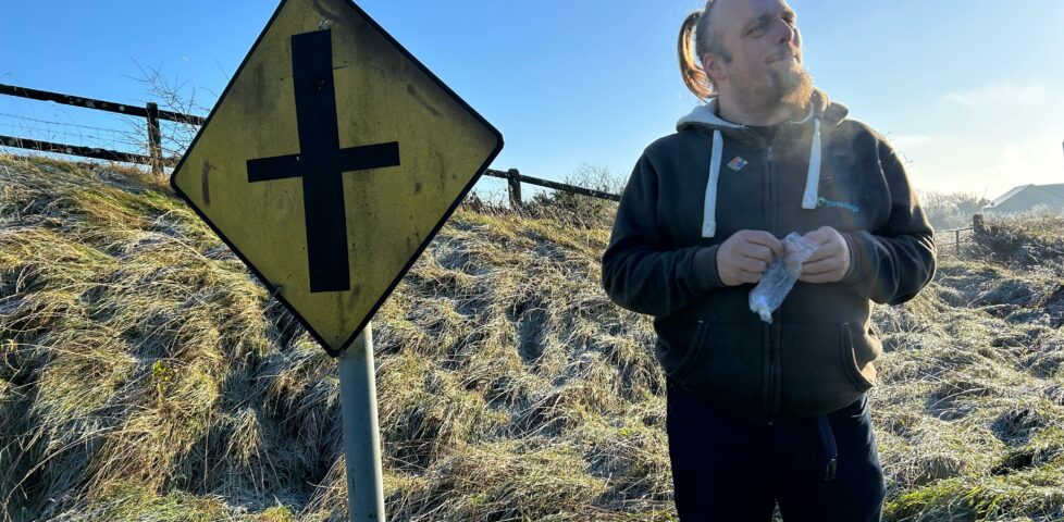 Dan smiles as he opens a zip-lock bag containing a geocaching logbook, alongside a diamond-shaped Irish 'crossroads' road sign.