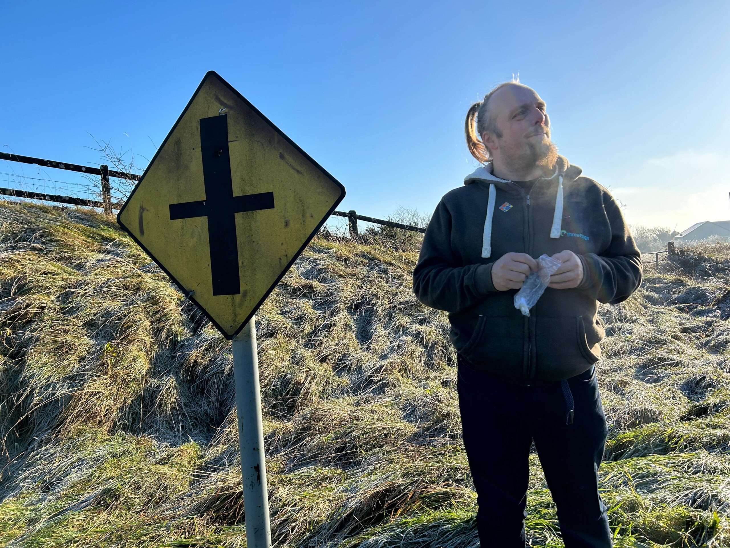 Full image of Dan smiles as he opens a zip-lock bag containing a geocaching logbook, alongside a diamond-shaped Irish 'crossroads' road sign.