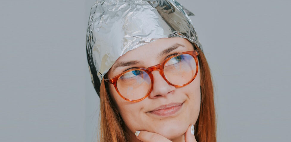 A white woman wearing glasses and a tinfoil hat touches her chin and looks upward, as if deep in thought.