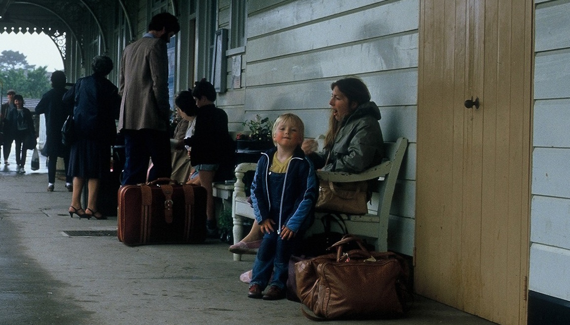 Full image of Dan, aged ~4, stands on a railway station platform alongside his mother, yawning, on a bench. It is overcast and drizzly.