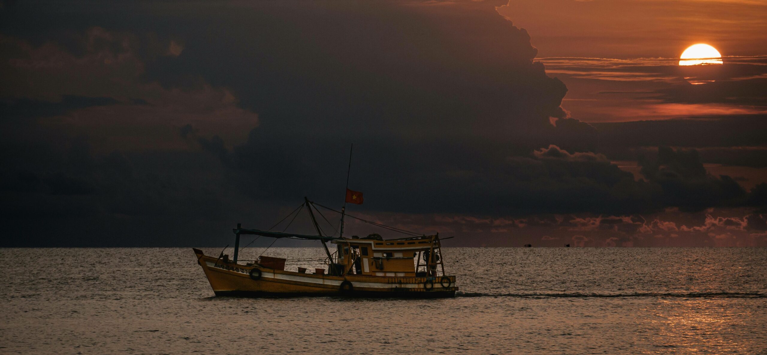 Full image of As the sun sets behind growing clouds, a small fishing vessel flying a red flag glides across a moderately-smooth ocean.