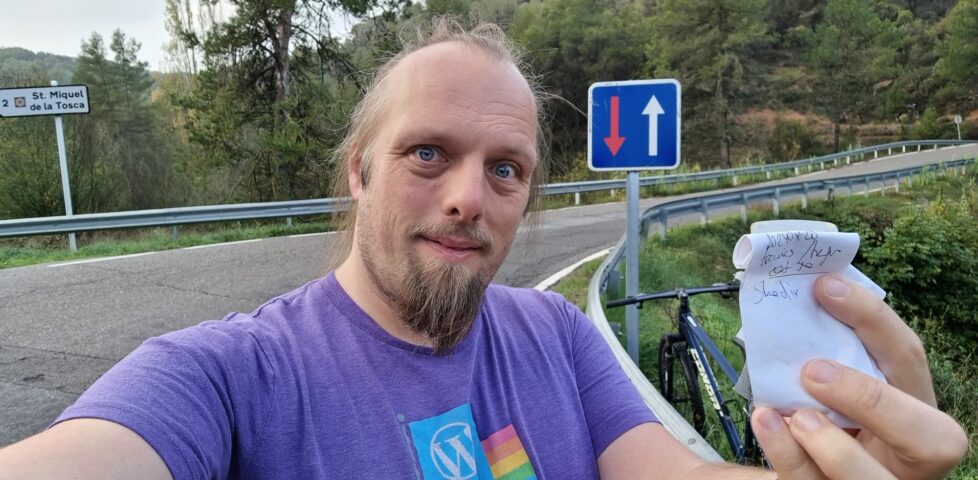 Dan, holding a small geocache logbook. A bike and a road bridge can be seen in the background.