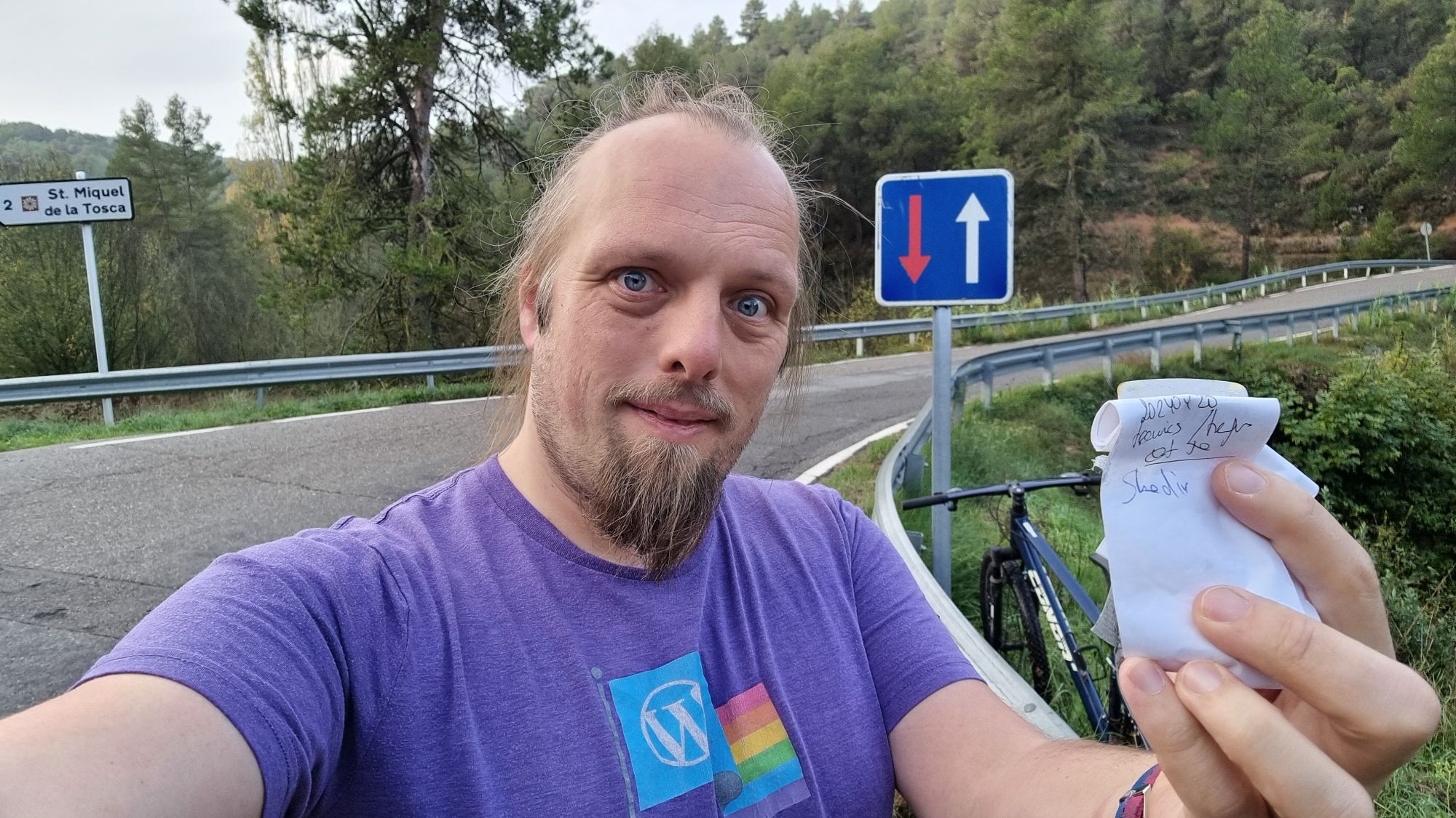Full image of Dan, holding a small geocache logbook. A bike and a road bridge can be seen in the background.