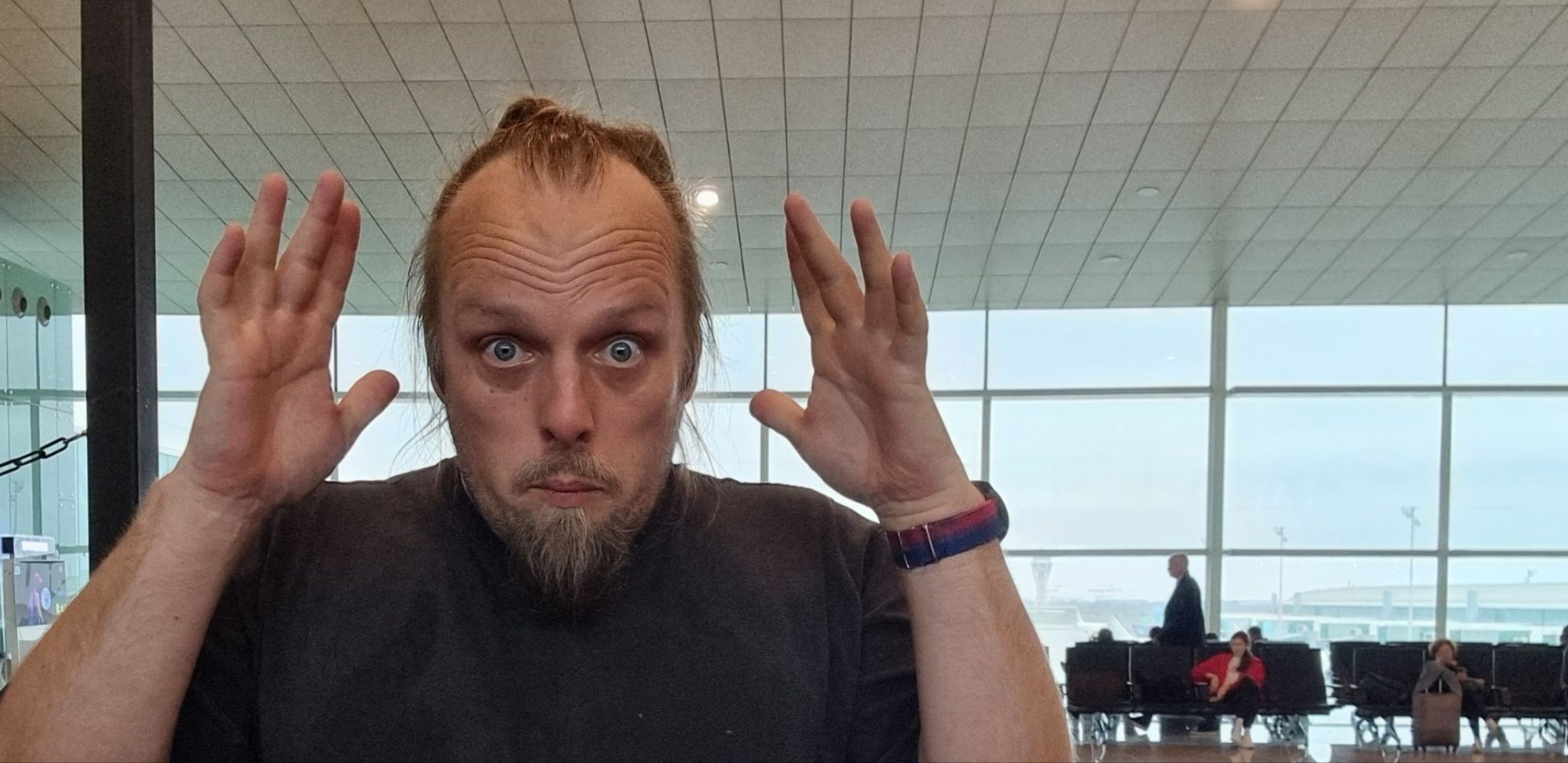 Full image of Dan, standing in an airport departure lounge, mimes "mind blown" to the camera.