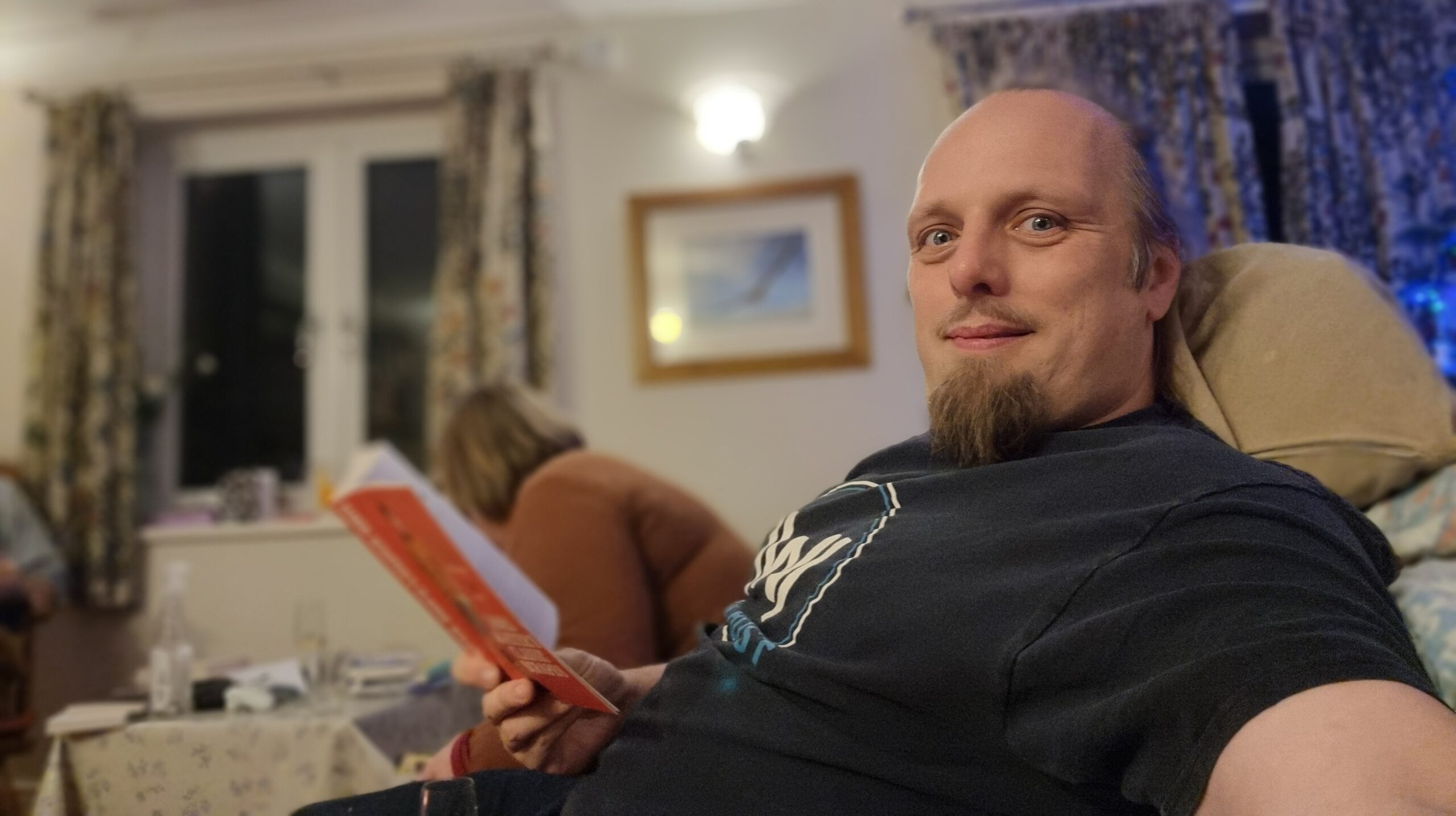 Full image of Dan sits by firelight reading a red-spined book.