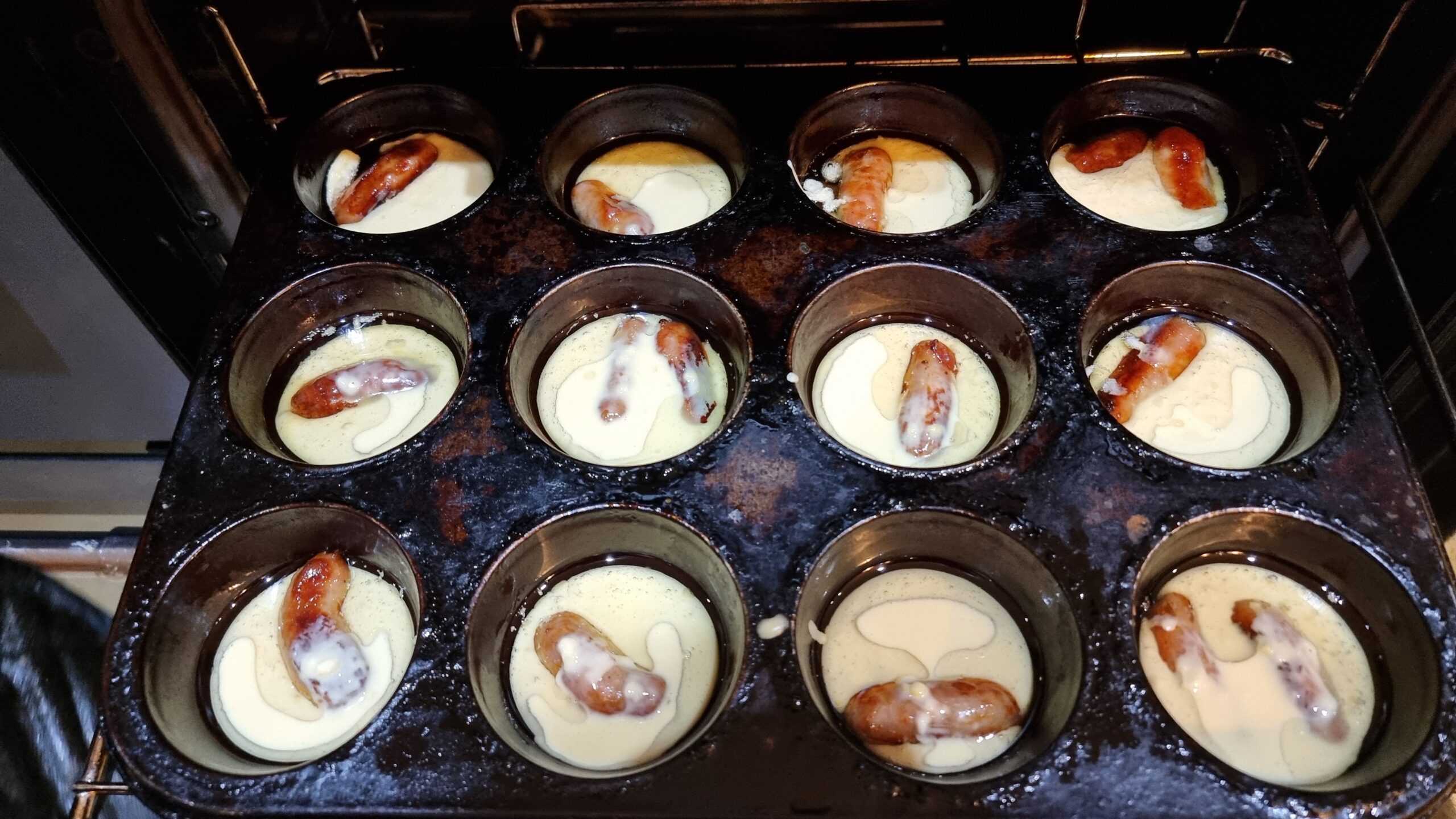 Full image of Cupcake-sized Yorkshire pudding batter cups, each with a cocktail sausage or two inside, being inserted into an oven.