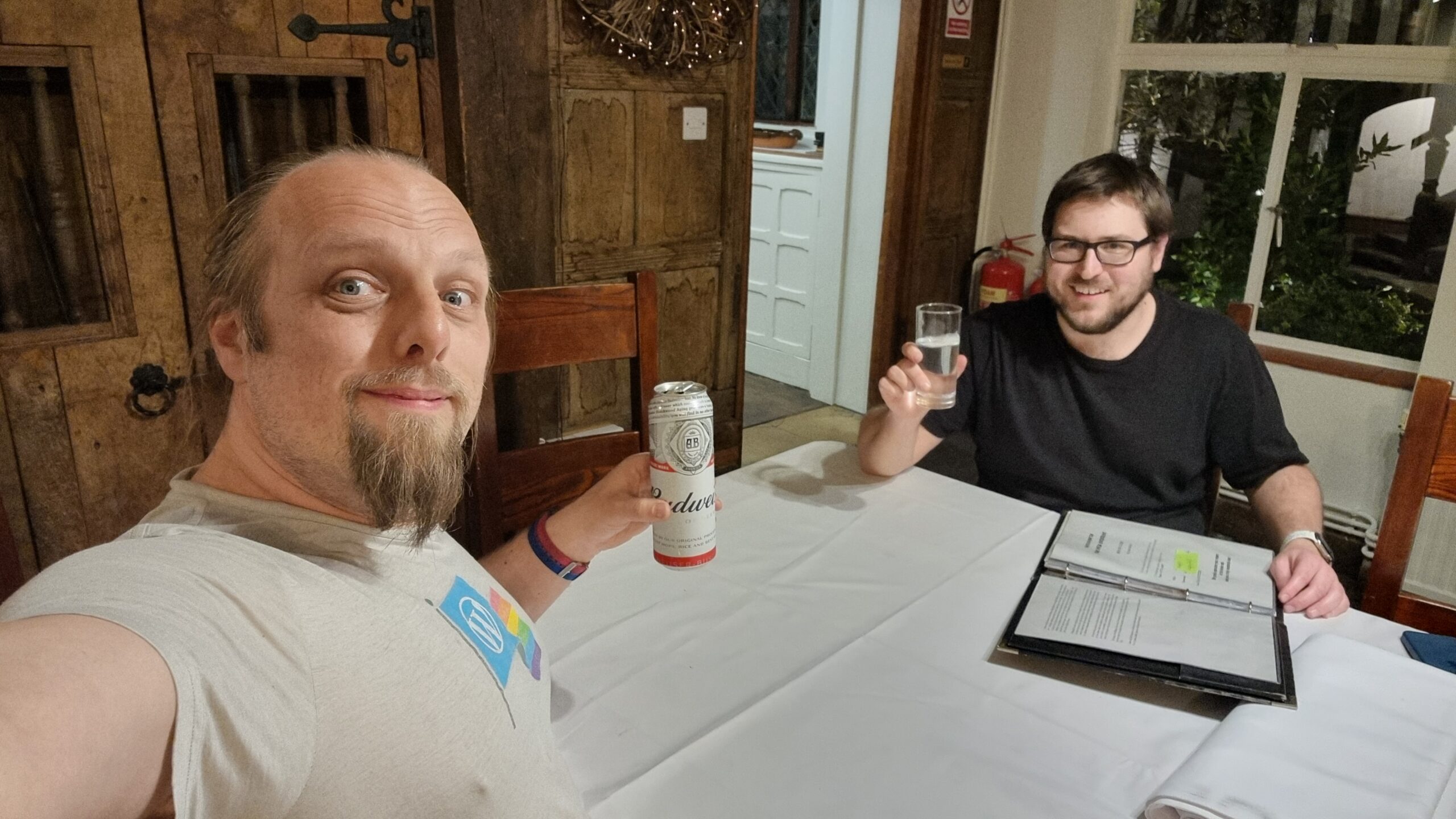 Full image of Dan, dressed in white, half-smiling, raises a can of Budweiser beer as he sits alongside another man, dressed in black, raising a glass of a clear liquid.
