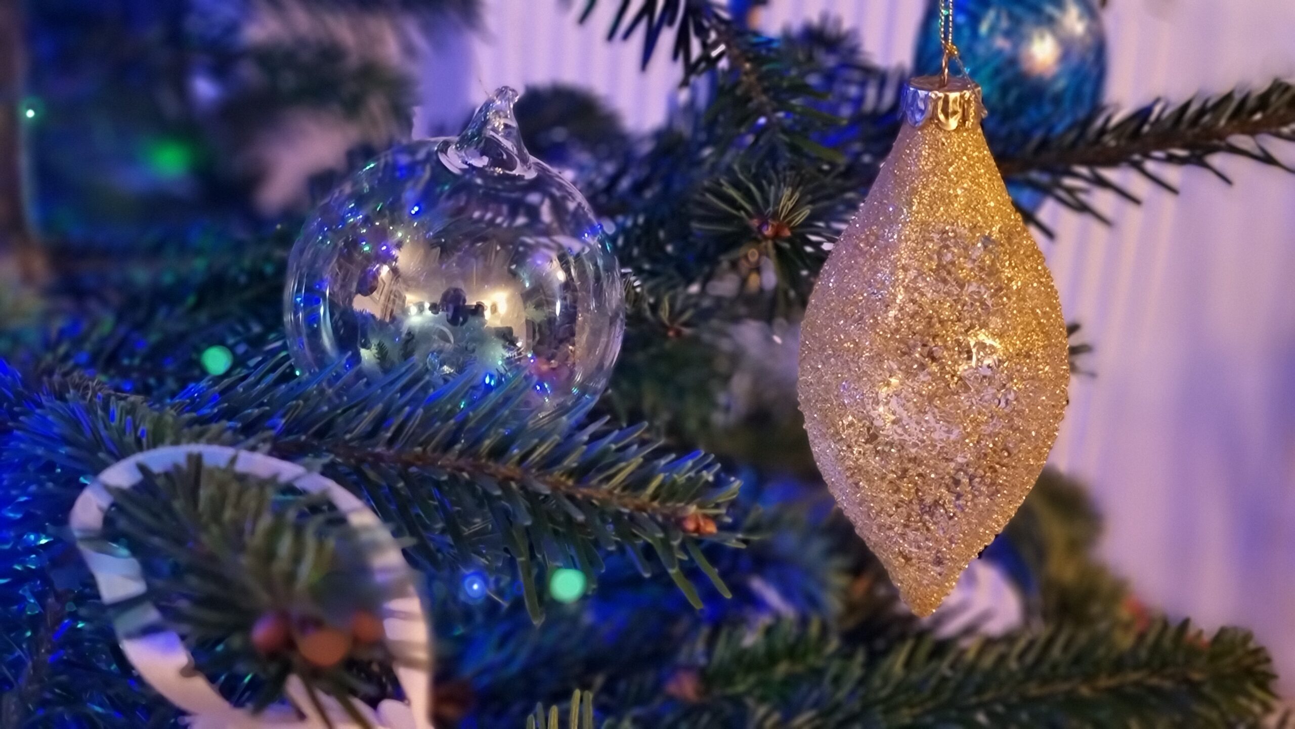 Full image of A highly-reflective 'soap bubble' glass bauble hangs alongside a glittery gold teardrop-shaped bauble, lit by green and blue fairy lights.