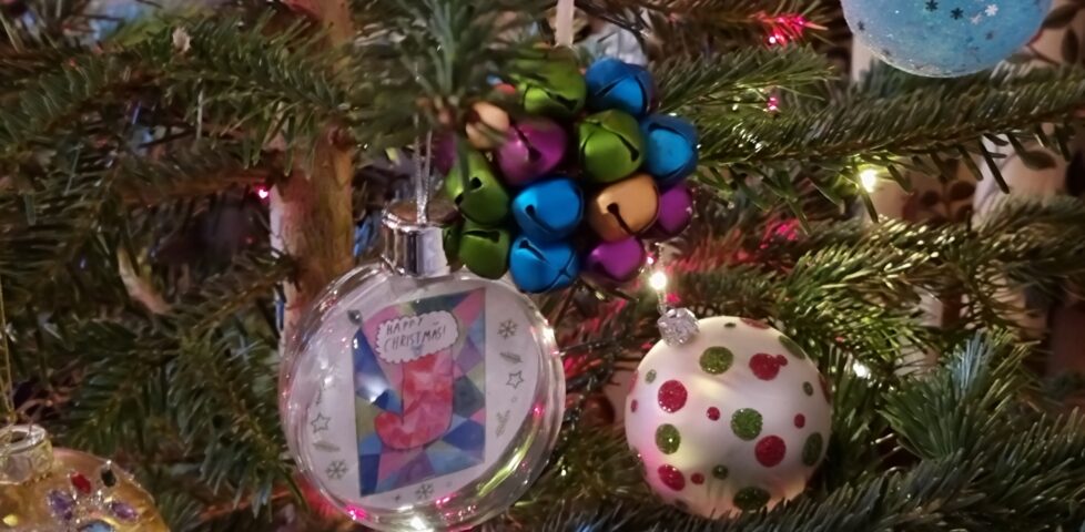A cluster of three baubles hangs among pink and white fairy lights: one is a multicoloured assortment of bells, another is a plain white bauble decorated with glittery green and red spots, a third is a transparent plastic sphere containing a colourful children's drawing of a stocking.