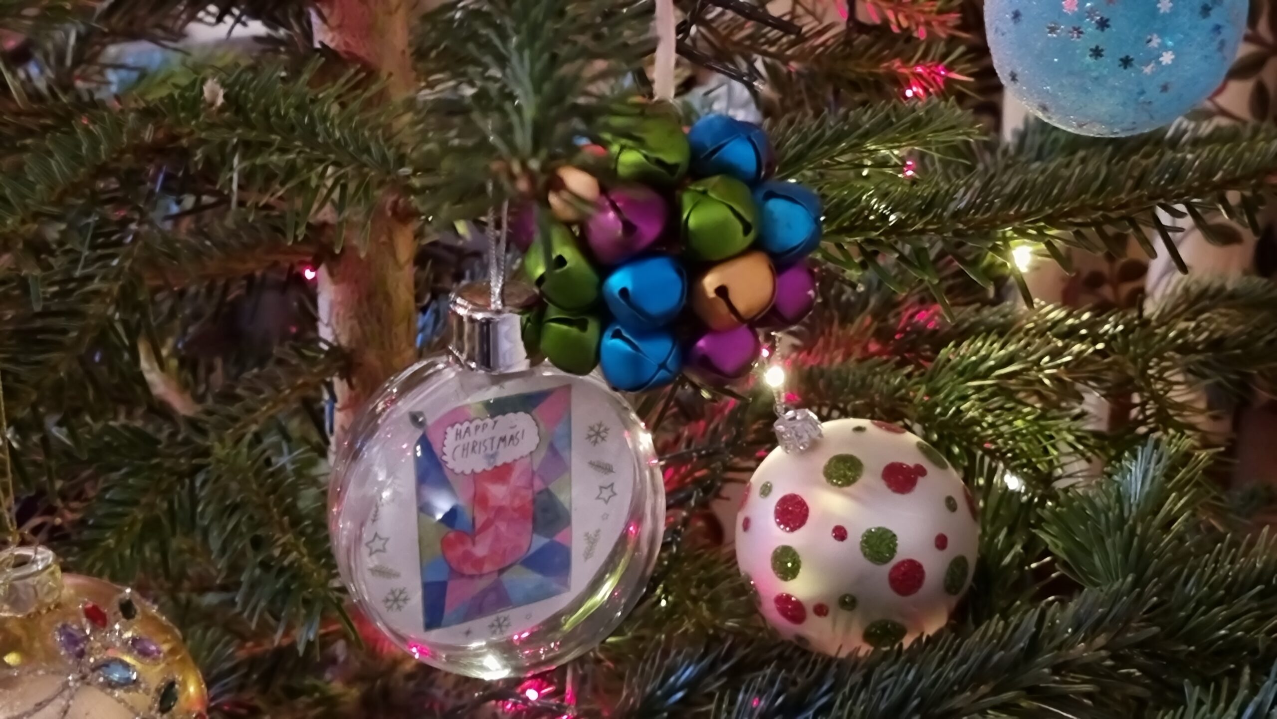 Full image of A cluster of three baubles hangs among pink and white fairy lights: one is a multicoloured assortment of bells, another is a plain white bauble decorated with glittery green and red spots, a third is a transparent plastic sphere containing a colourful children's drawing of a stocking.