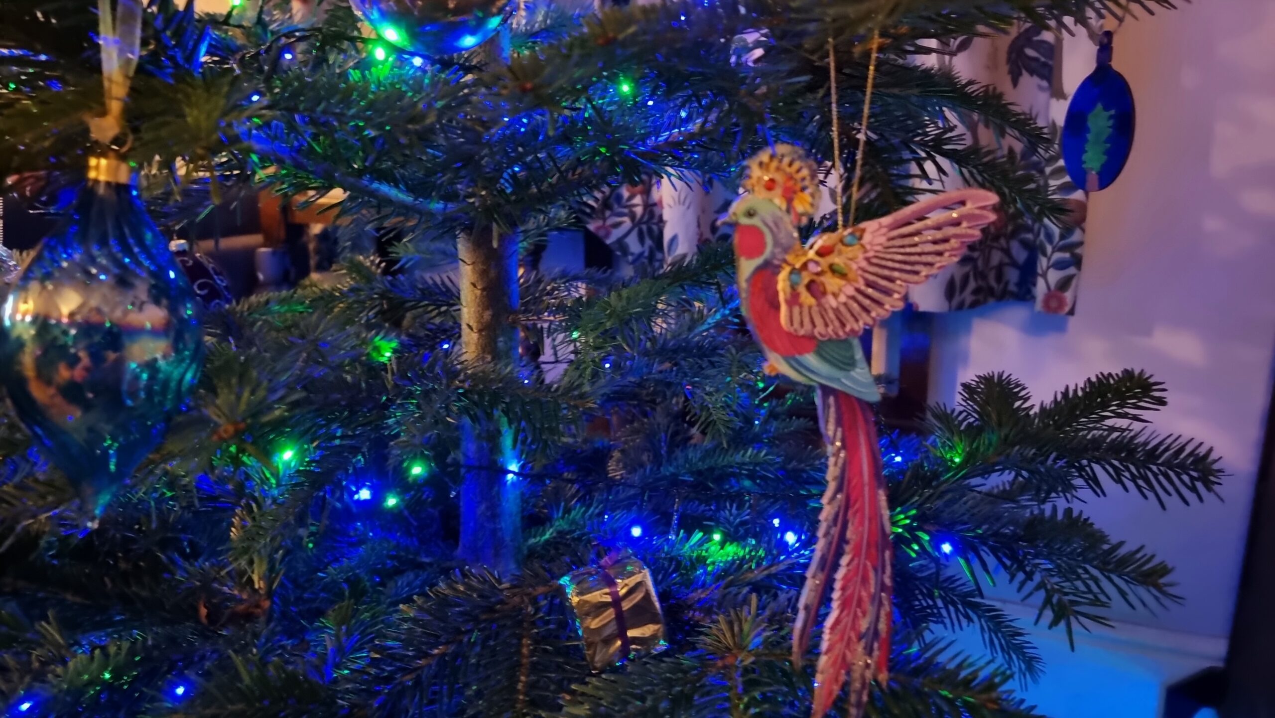Full image of A decoration in the form of a bejewelled exotic bird hangs between a traditional bauble with a rippled texture and a hand-painted decoration showing a potted tree.