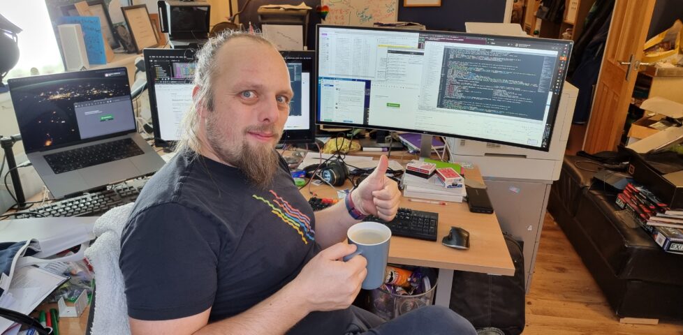 Dan, a white man with a goatee-style beard and long hair tied back, wearing a black t-shirt with an abstract pride rainbow, holds a mug of coffee in one hand and gives a thumbs-up with the other. Behind him, three computer screens show a mixture of code, source control, chat, email, and web browser windows. His desk is cluttered.