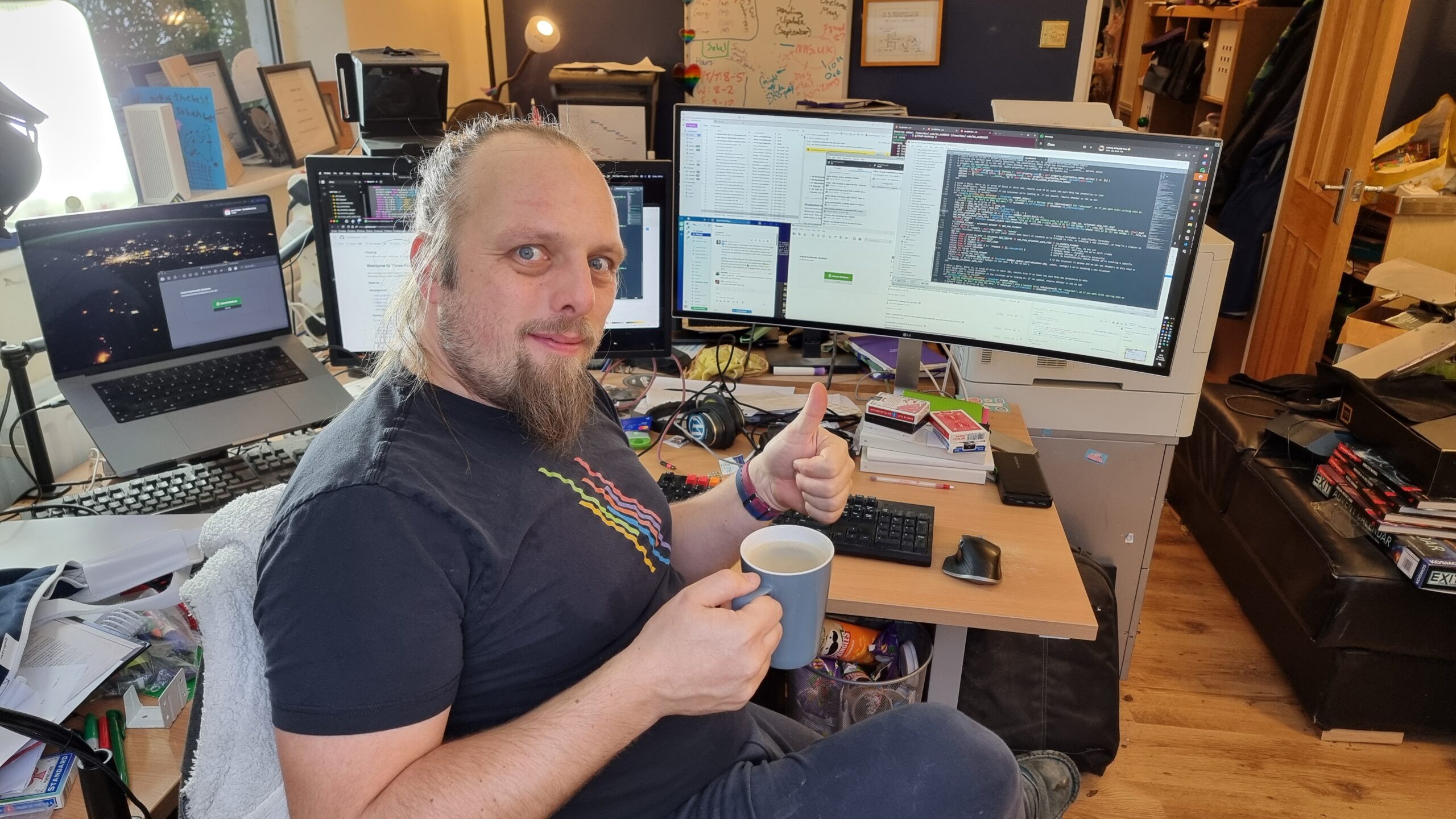 Full image of Dan, a white man with a goatee-style beard and long hair tied back, wearing a black t-shirt with an abstract pride rainbow, holds a mug of coffee in one hand and gives a thumbs-up with the other. Behind him, three computer screens show a mixture of code, source control, chat, email, and web browser windows. His desk is cluttered.