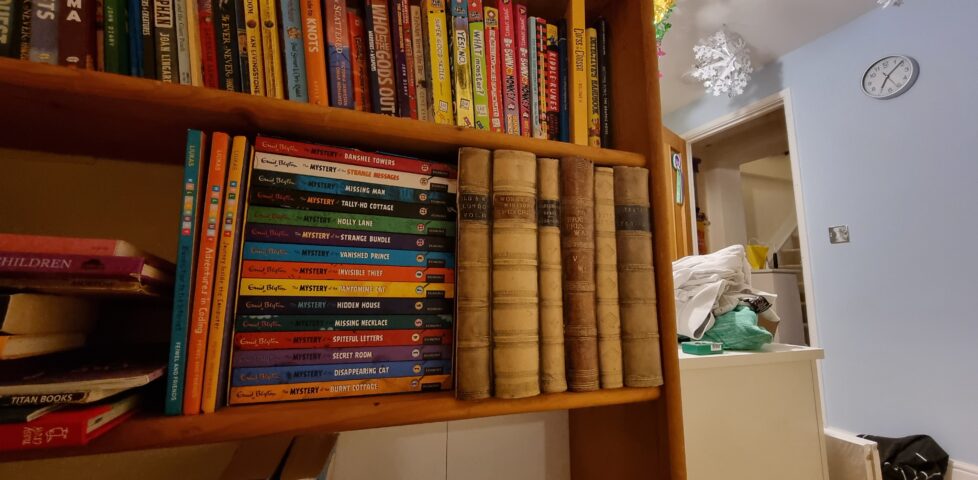 A bookcase in a child's bedroom, with Christmas decorations visible in the background. Several very old looking books stand conspicuously on the shelf.