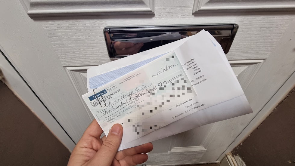 Full image of In front of a letterbox, an hand holds an open envelope and a (censored) Danske Bank cheque for £219.
