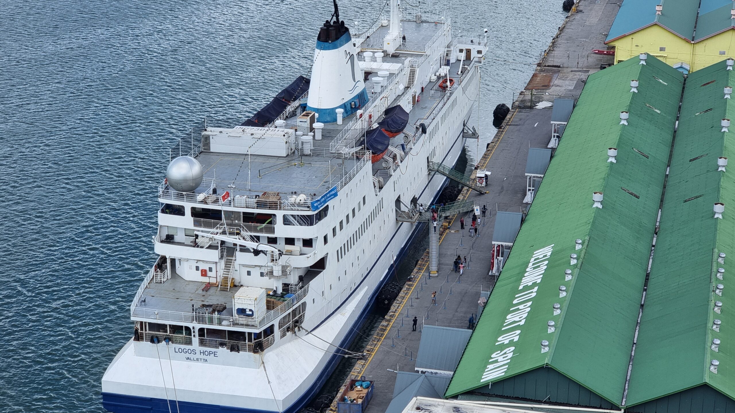 Full image of White and blue passenger ship docked alongside a building whose roof reads 'Welcome to Port of Spain'.