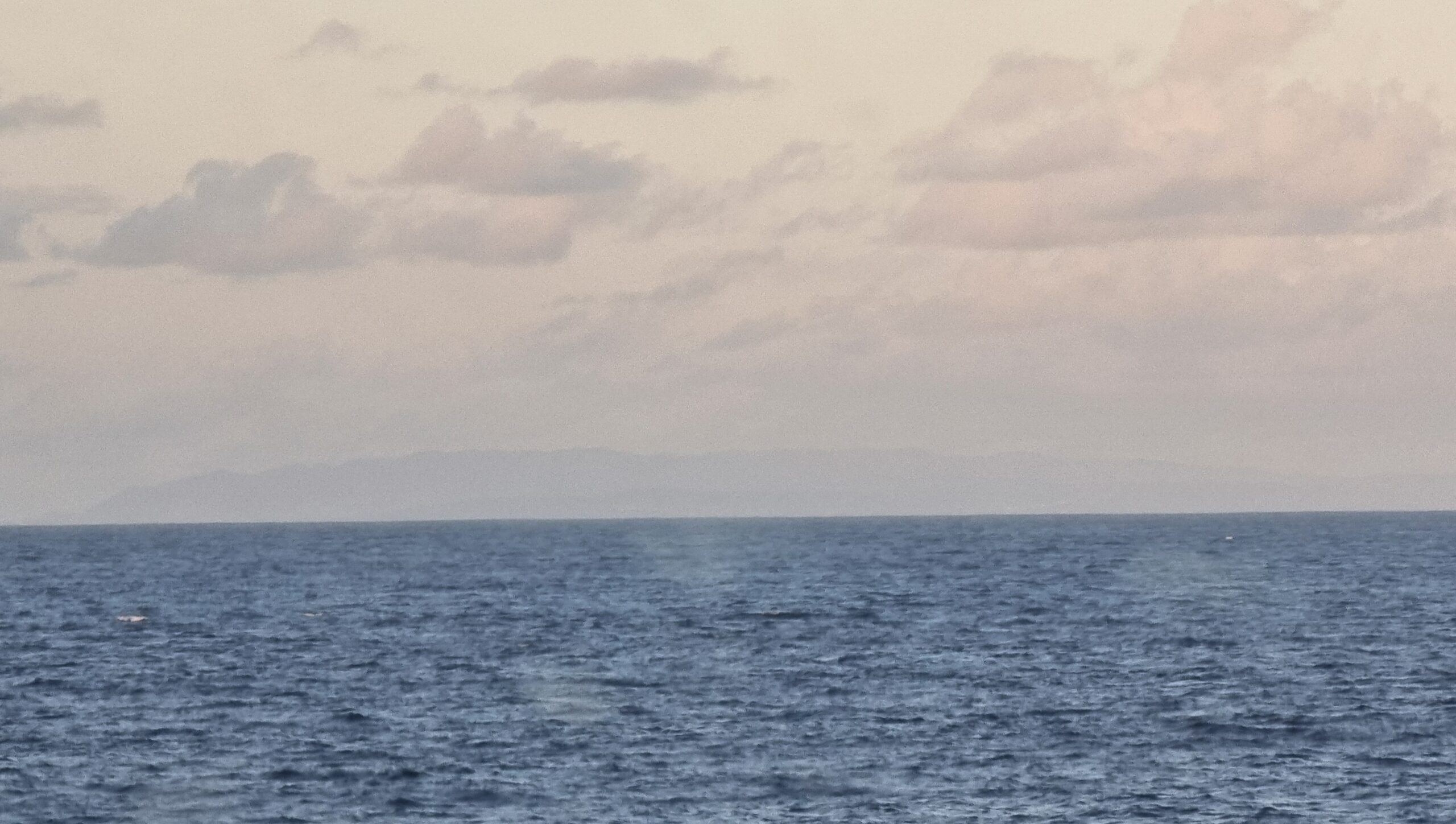 Full image of Across the sea, a rugged island begins to emerge over the horizon, through distant clouds.