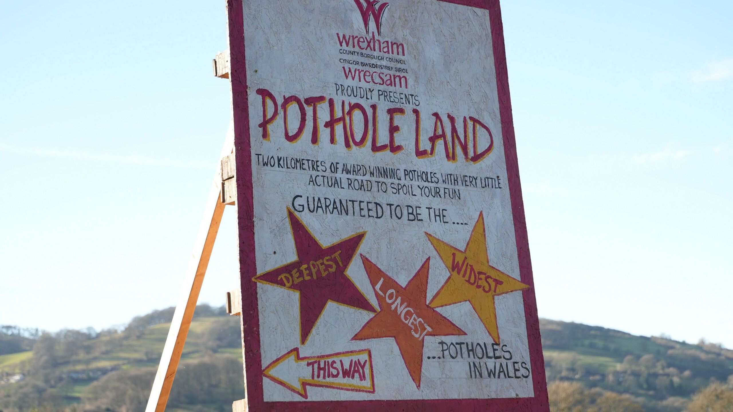 Full image of Humorous handmade road sign welcoming visitors to Wrexham as 'Pothole Land', stating that there are 'two kilometres of award-winning potholes with very little actual road to spoil your fun' and guaranteeing 'the deepest, longest, vs widest potholes in Wales'.