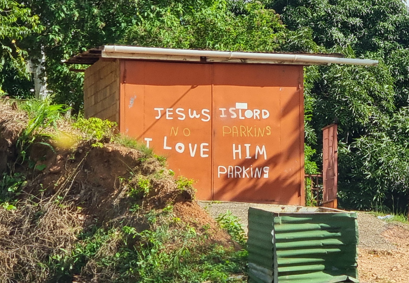 Full image of Orange garage on a tropical hillside, on the front of which are painted the words: 'Jesus is Lord. I love him.' And then below and between those lines: 'No parking'.