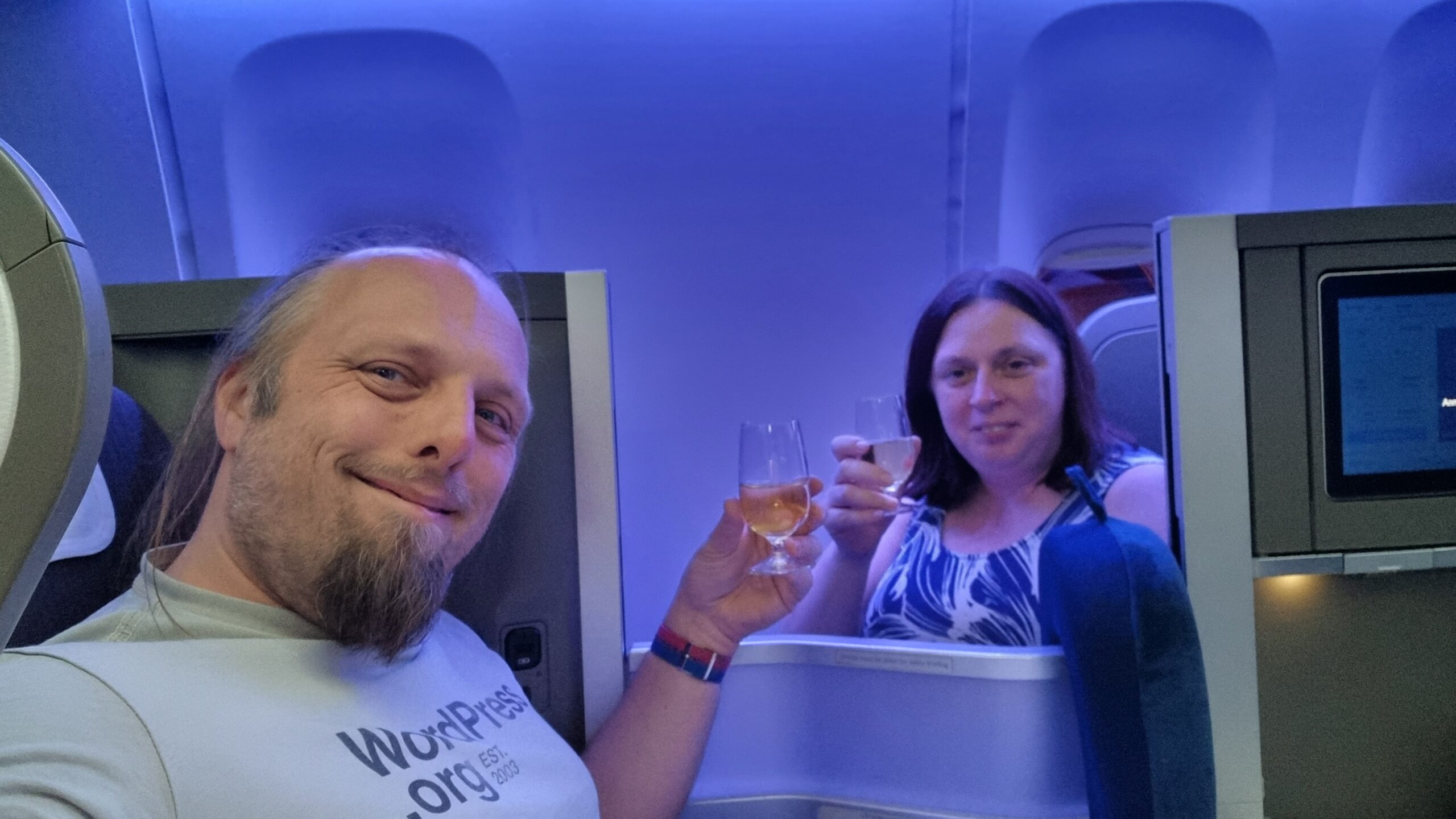 Full image of Dan and Ruth clink champagne glasses between the booths of their club class seats.