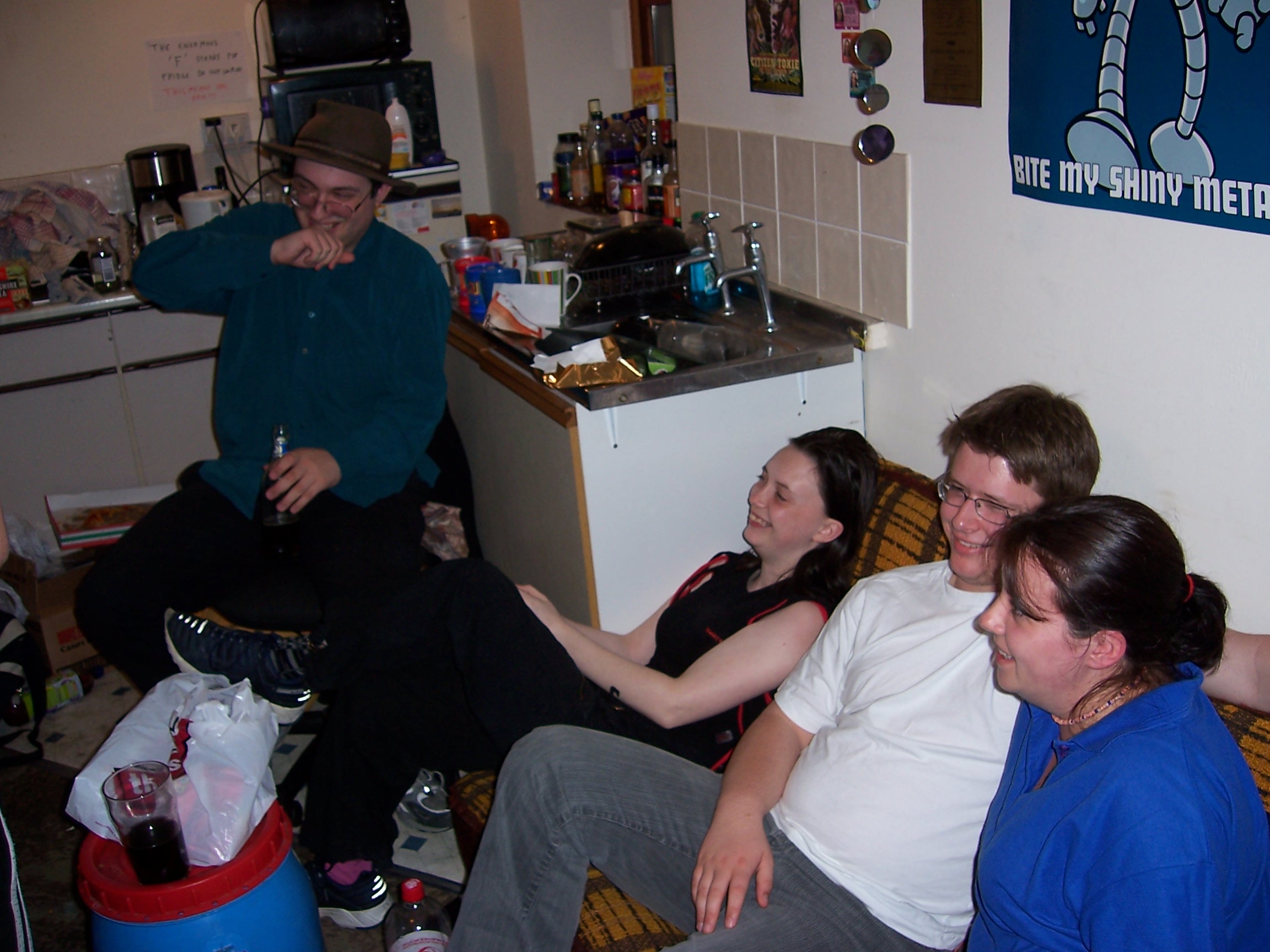 Full image of Four young people, smiling in laughing, sit in a cluttered and messy flat.