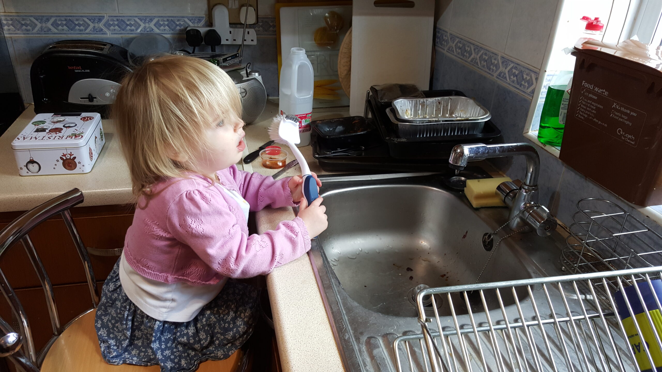Full image of A toddler with long blonde hair, wearing a pink cardigan, sits on a tall stool in front of a kitchen sink, holding a long-handled scrubbing brush.