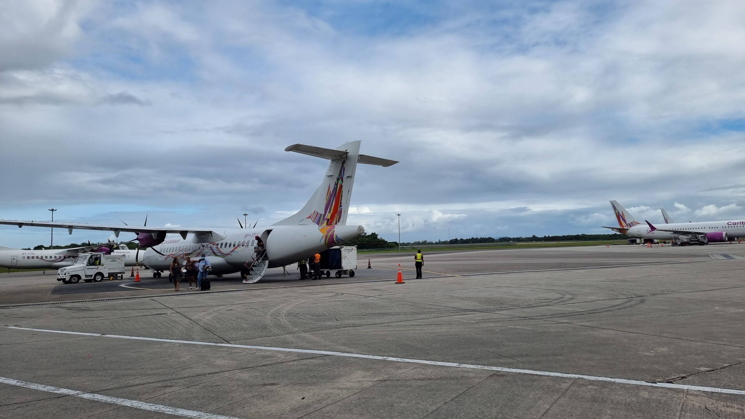 Full image of Medium-sized two-engine turboprop aircraft on tarmac runway, facing away from the photographer, under slightly cloudy blue skies. Caribbean Airlines livery (colourful hummingbird on tail).