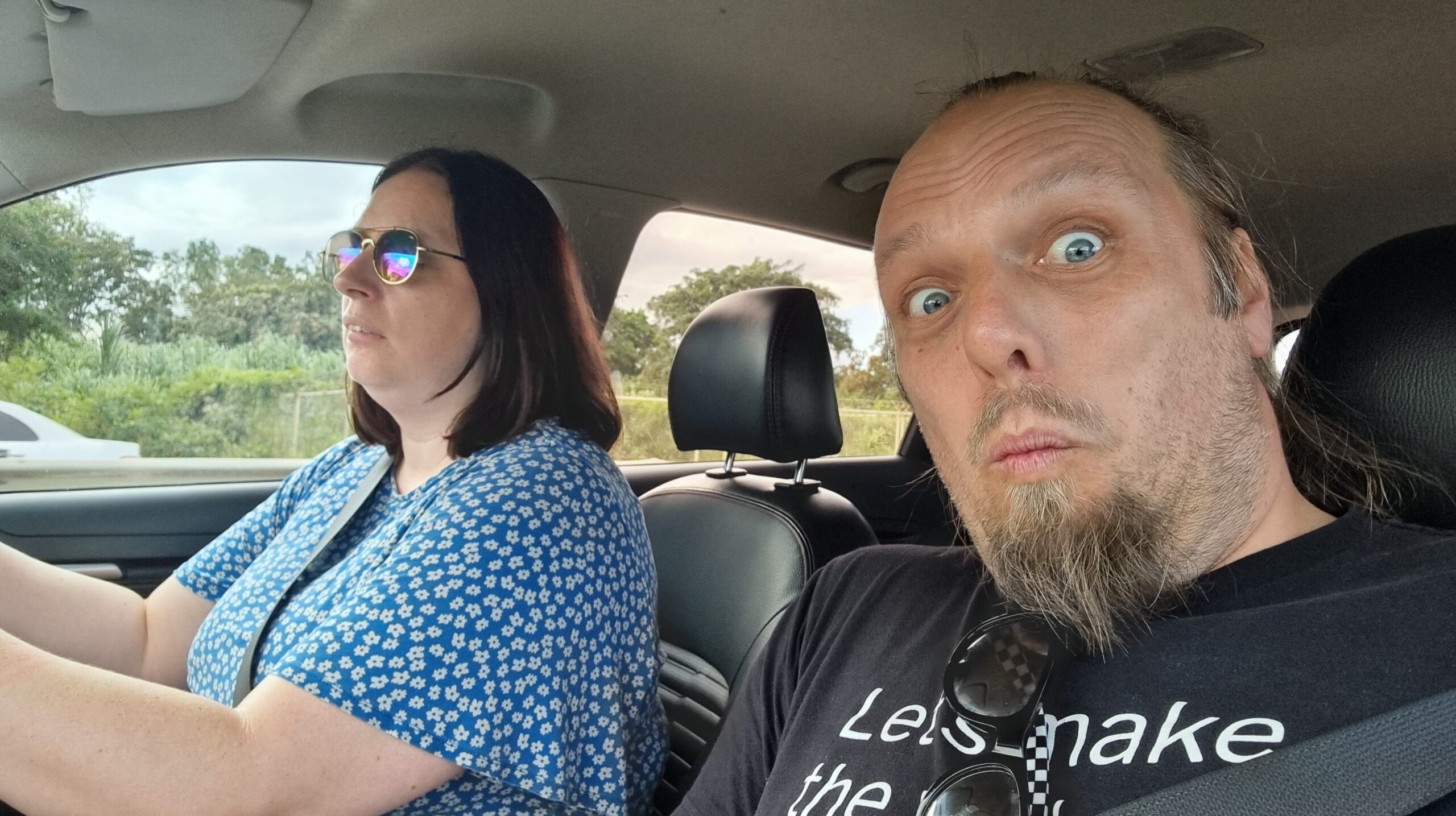 Full image of Ruth drives a car; Dan, in the passenger seat, looks into the camera as he takes a selfie.