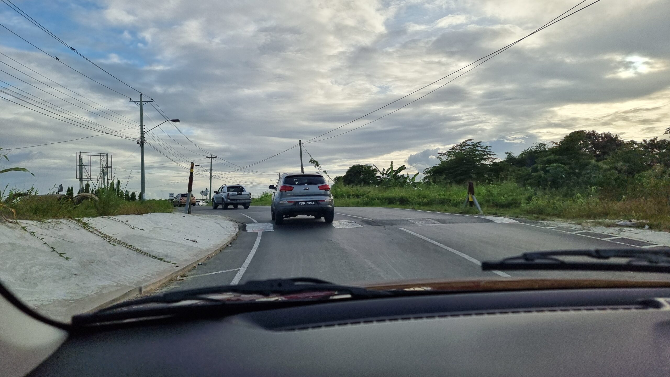 Full image of View through a windscreen showing cars ahead going over a speed bump and winding along a road between tropical shrubbery, with power lines overhead.