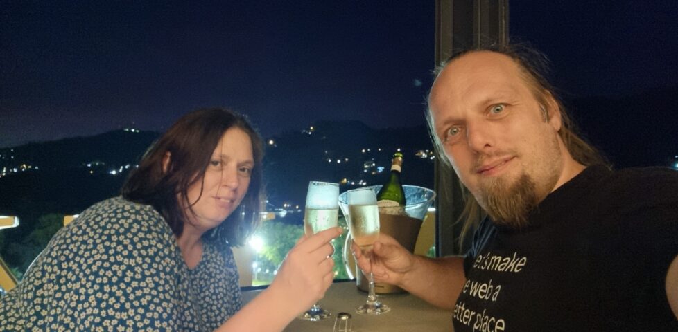 Dan and Ruth clink champagne flutes under a dark sky, with city lights visible below them.