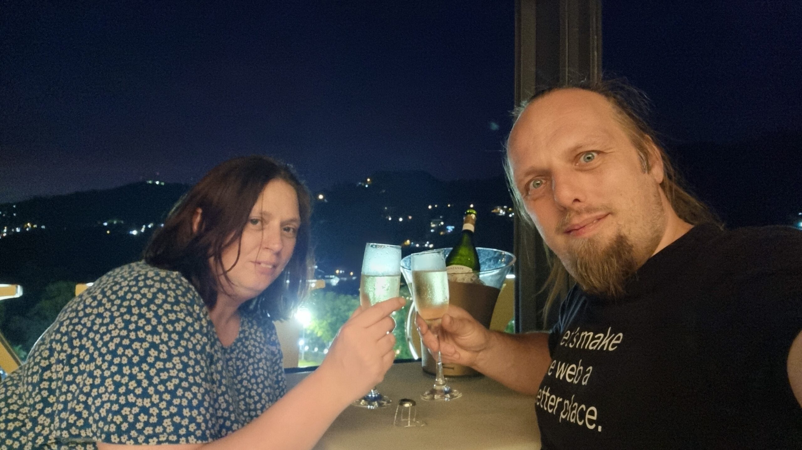 Full image of Dan and Ruth clink champagne flutes under a dark sky, with city lights visible below them.