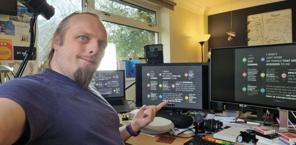 Dan, wearing a purple t-shirt, looks at the camera while pointing at the centre of three computer screens which share a cluttered desk, each of which shows a stylised image version of the Automattic Creed.