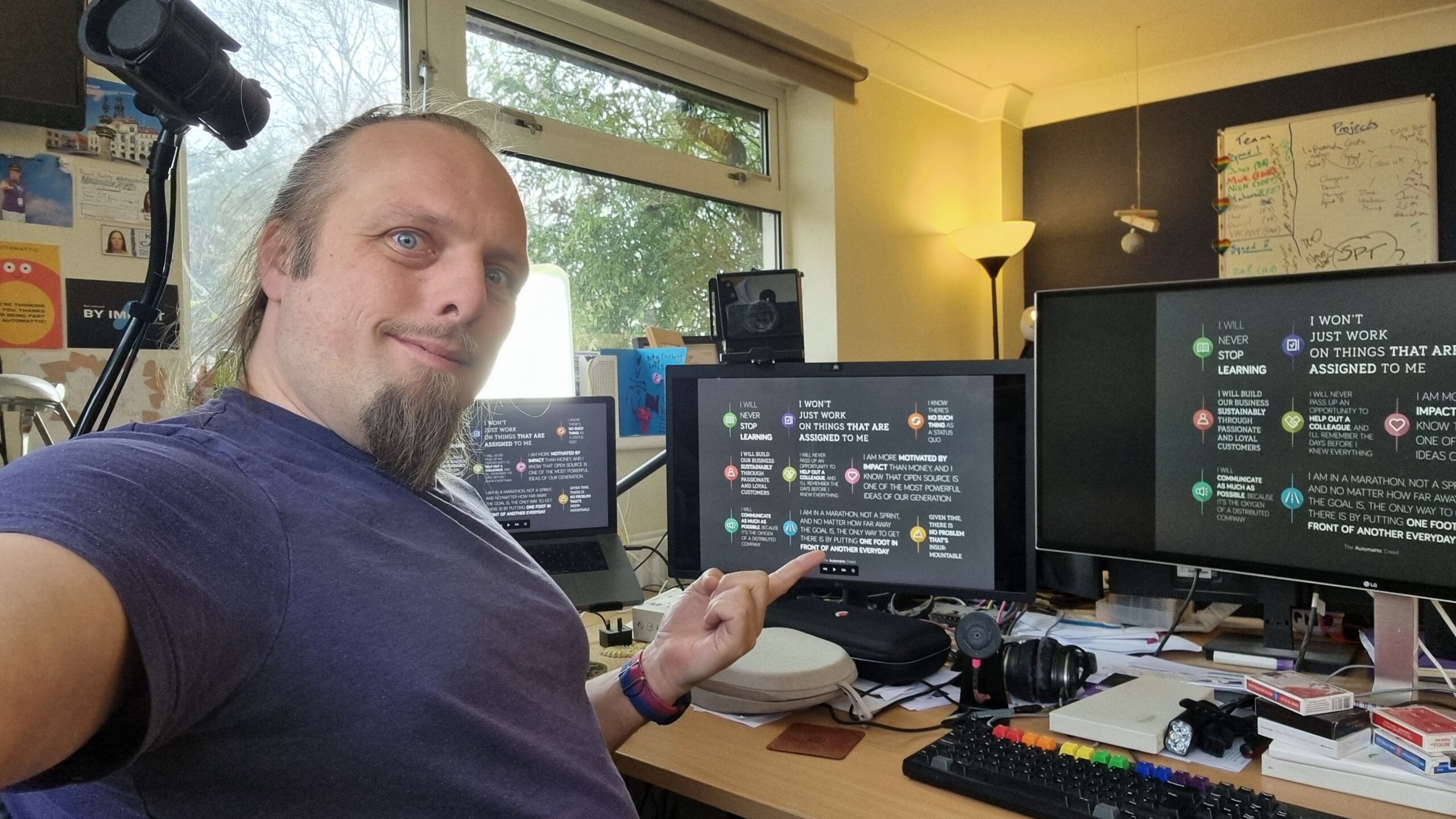 Full image of Dan, wearing a purple t-shirt, looks at the camera while pointing at the centre of three computer screens which share a cluttered desk, each of which shows a stylised image version of the Automattic Creed.