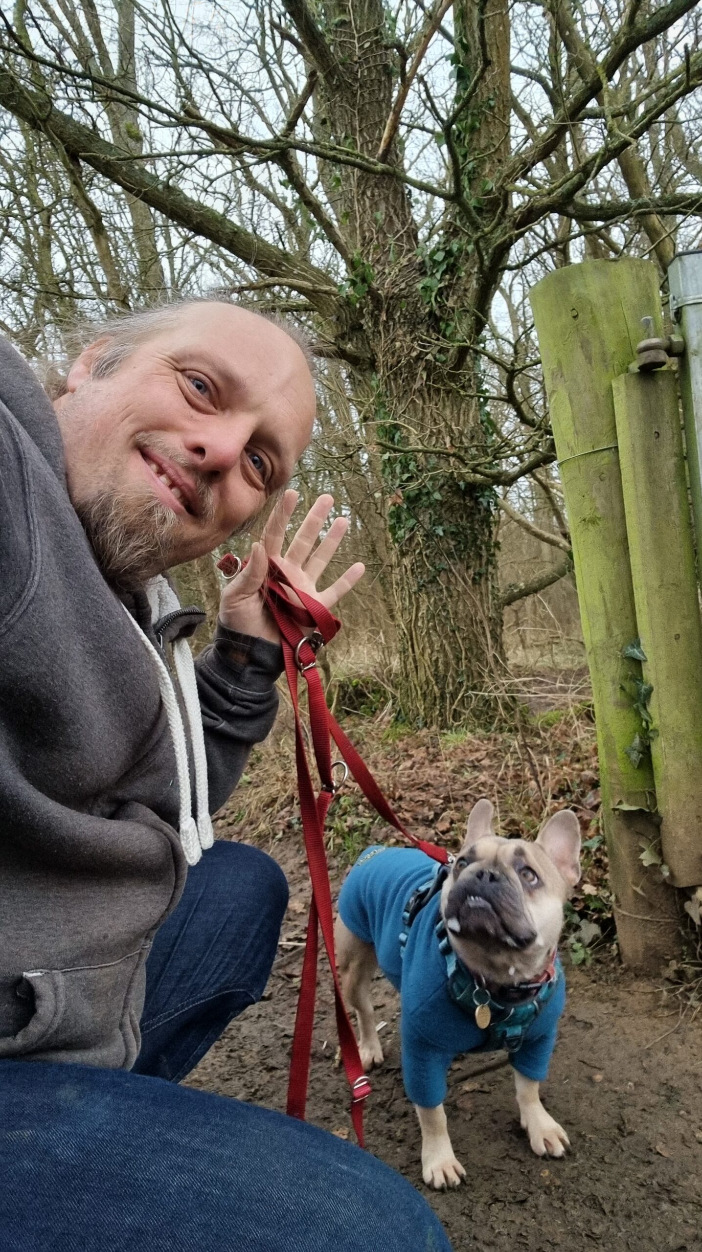 Full image of Dan, wearing a grey jacket at the edge of a wet and wintry forest, waves to the camera with a hand that also holds a dog lead, the other end of which is connected to a French Bulldog wearing a teal jumper.