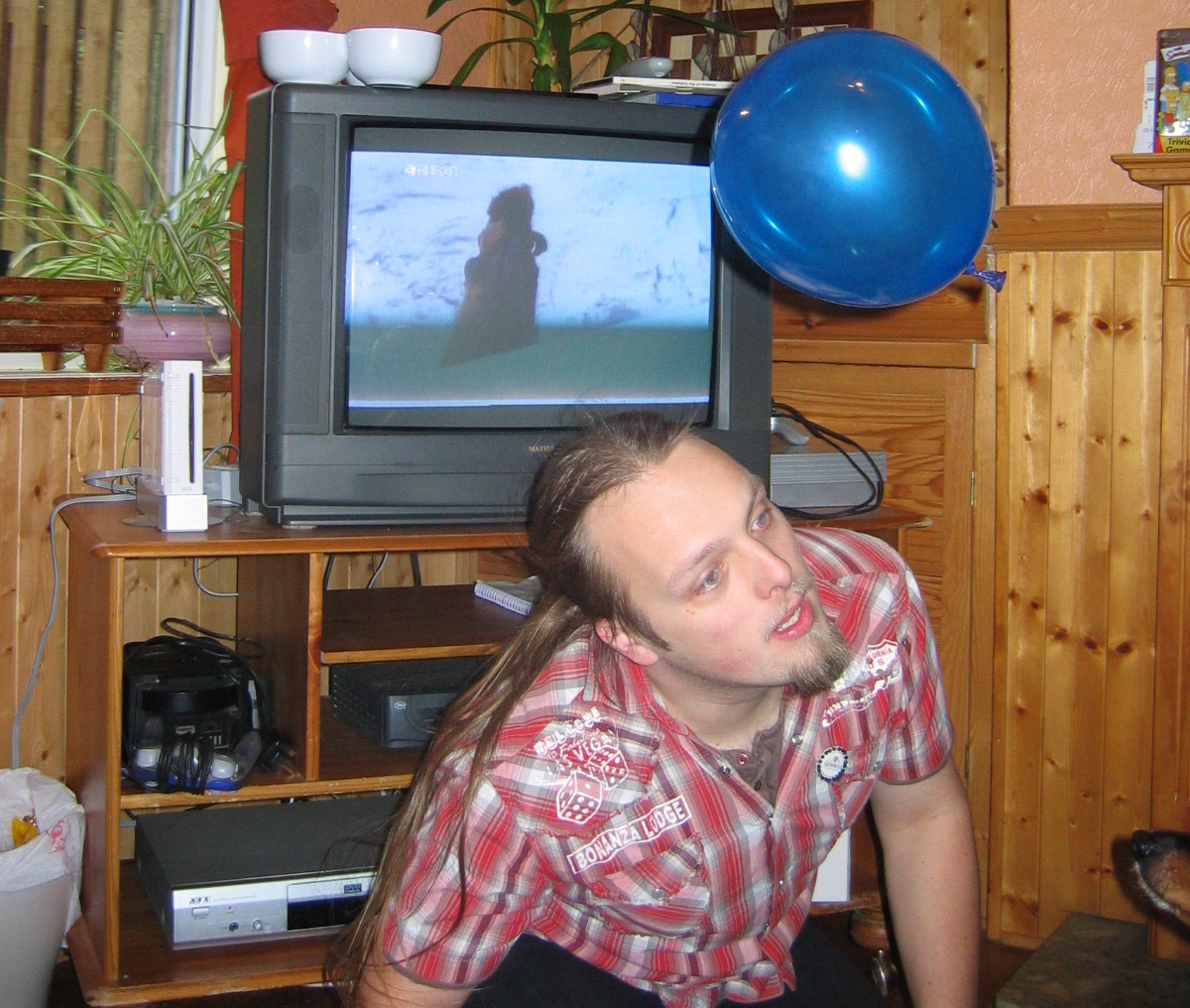 Full image of Dan, in his 20s, crouches awkwardly in front of a TV and a Nintendo Wii in a wood-panelled room as he attempts to headbutt a falling blue balloon.