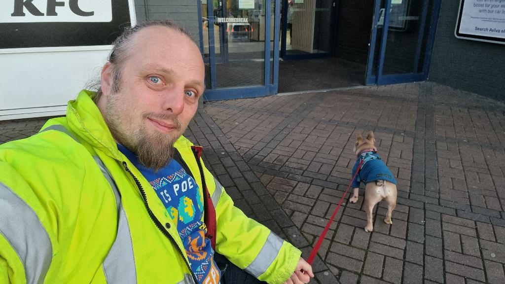 Full image of Dan, wearing a high-vis jacker, sits at a bench outside a motorway service station building. His dog, a small French Bulldog, pulls at her lead towards the entrance.