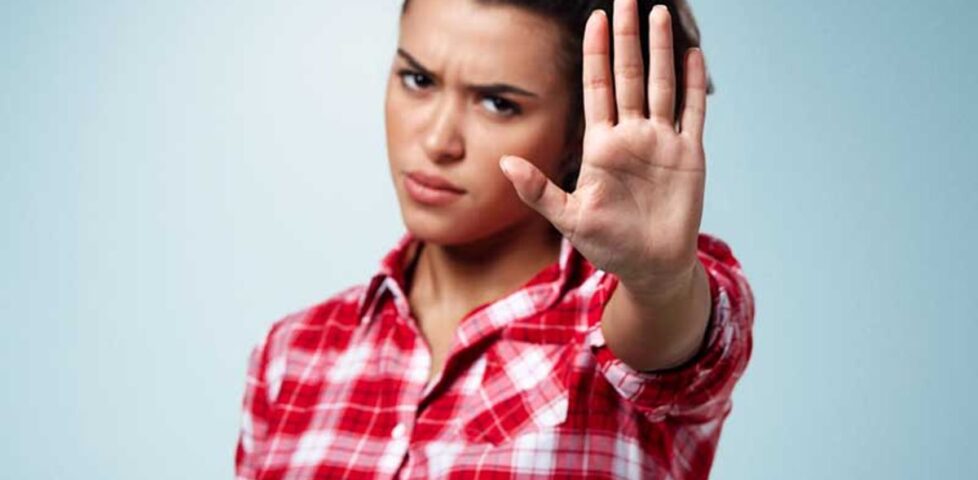 A white woman wearing a red-and-white flannel shirt frowns at the camera as she holds a hand up in a "halt" position.