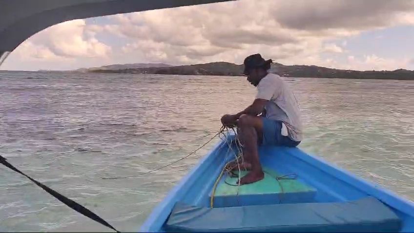 Full image of A man sits on the prow of the Cariad, over a sandbar, pulling in her anchor.