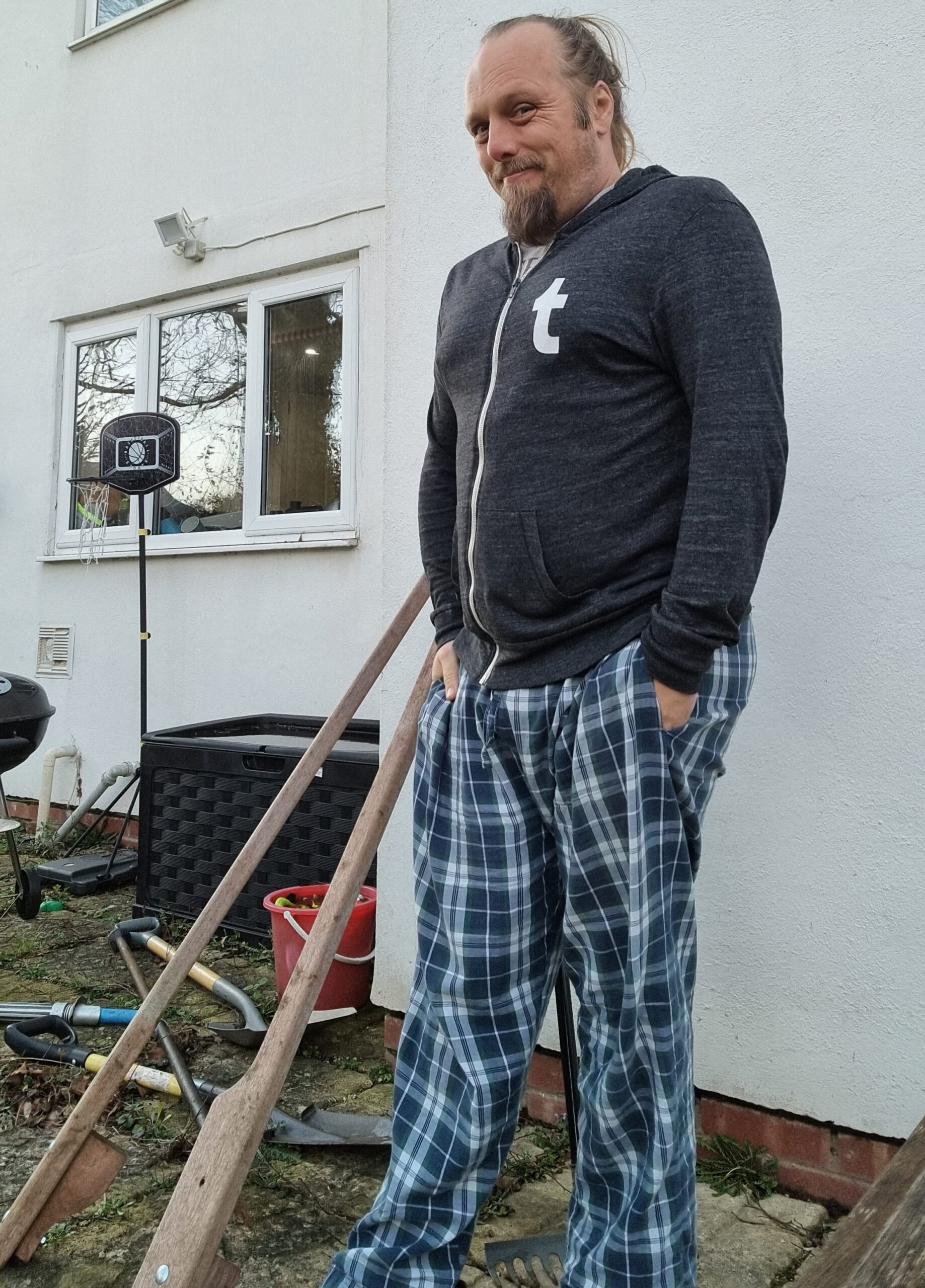 Full image of A white man stands outside a white house, looking cold. His hands are buried in the pockets of his checked pyjama bottoms, and his upper half is wrapped tightly in a Tumblr-branded grey jumper. Various childrens toys litter the garden around him.
