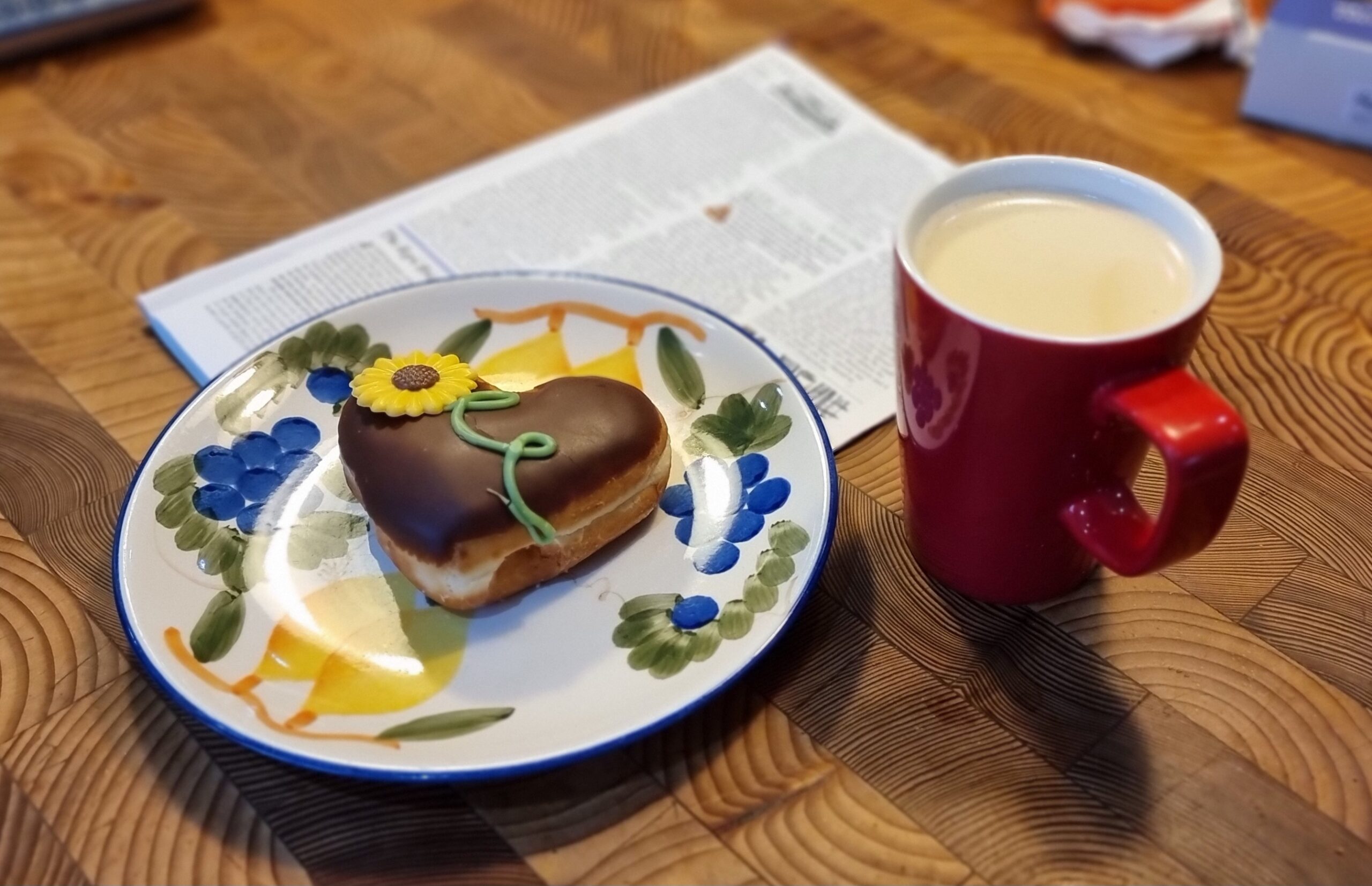Full image of A wooden kitchen surface containing a red mug full of freshly-brewed coffee alongside a plate painted with fruits on which sits a heart-shaped doughnut, topped with chocolate and decorated with an iced motif of a sunflower. Beneath the plate, out-of-focus, are the pages of a news periodical.