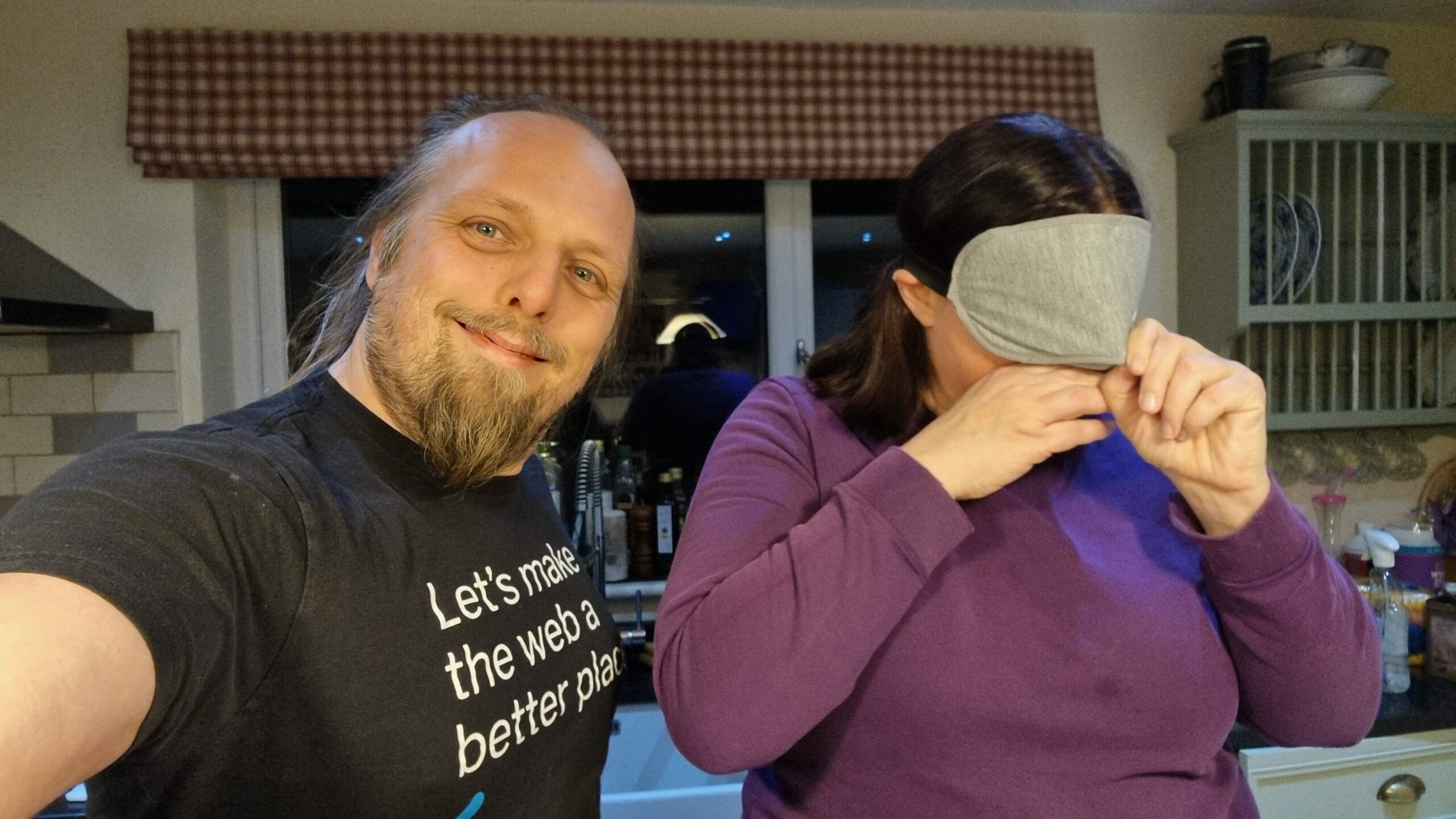 Full image of Dan, wearing a black t-shirt, smiles as he takes a selfie. Alongside him Ruth, wearing a purple jumper, adjusts a grey blindfold to cover her eyes.