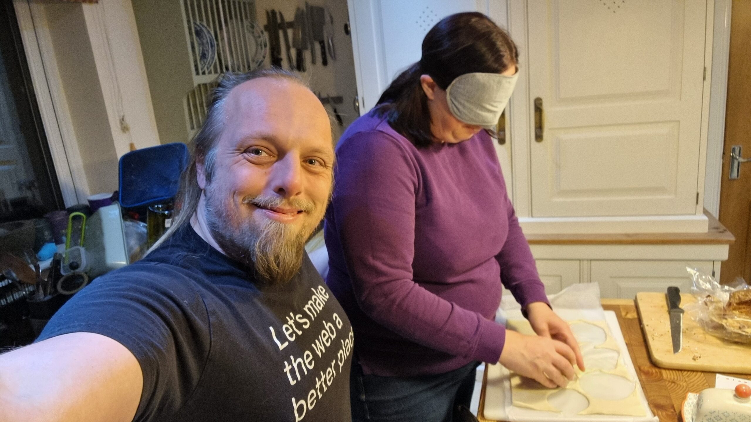 Full image of Dan takes a selfie showing himself, smiling, and Ruth, wearing a blindfold and balling up pastry on a wooden worksurface.