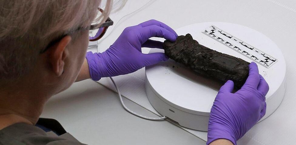 A researcher wearing purple sterile gloves carefully places a charred rolled scroll onto a white panel alongside a ruler, for x-ray photography purposes.