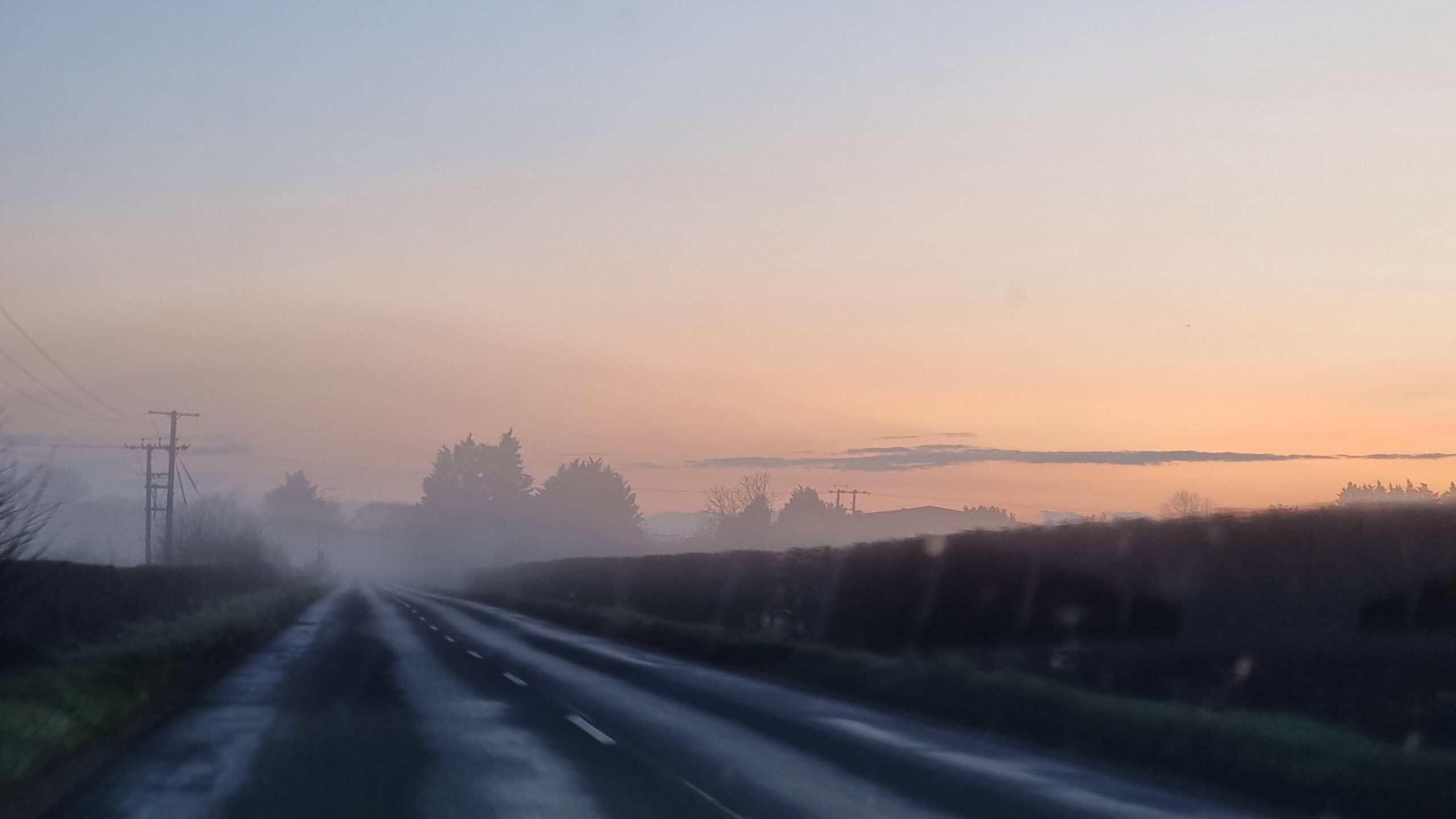 Full image of A wet rural road, lit by the sun's first light as it rises ahead, it's flanked by fields full of freezing fog.
