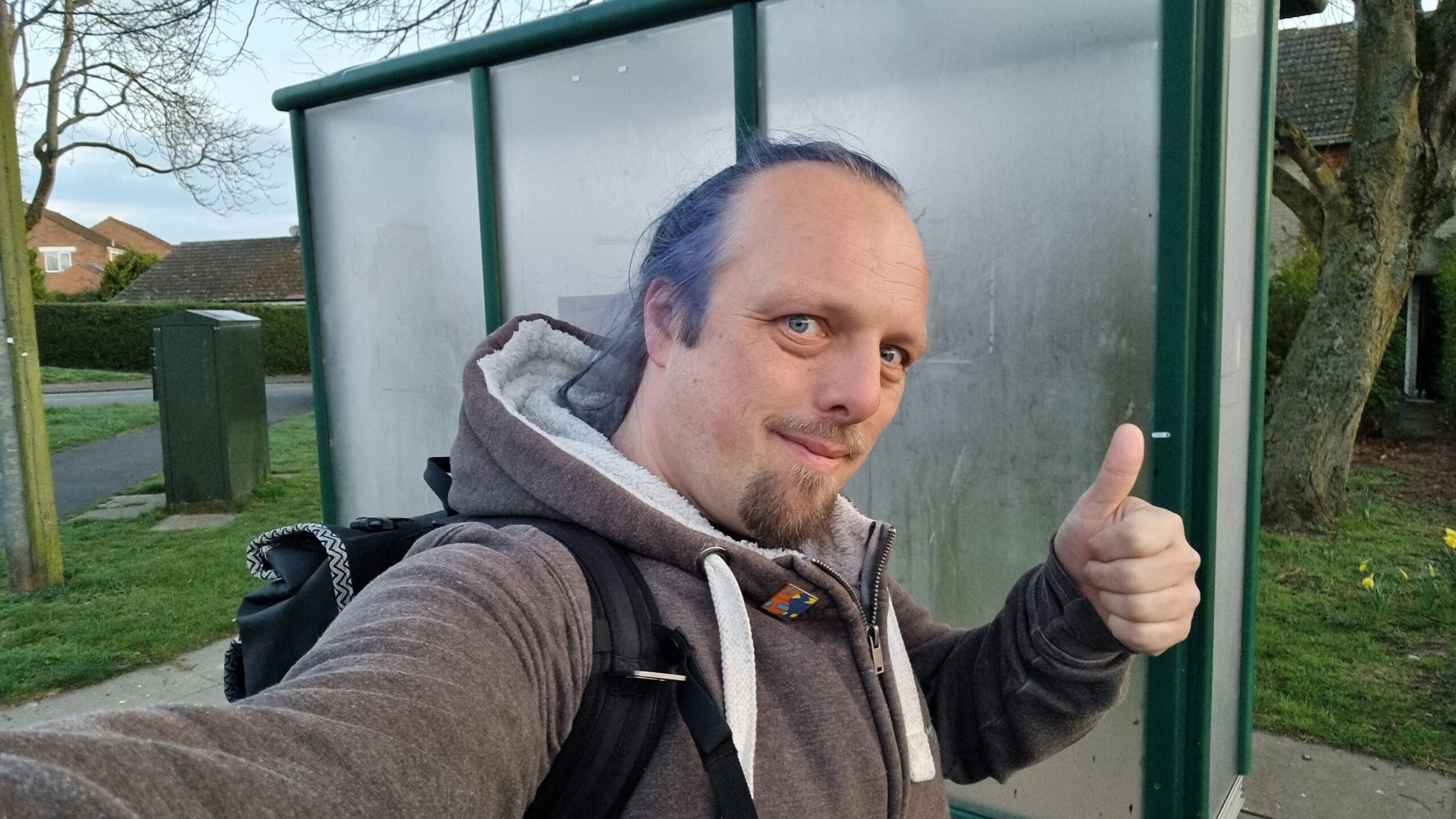 Full image of Dan, a white man with a trimmed goatee beard and dyed-blue hair tied back in a ponytail, stands alongside a suburban bus shelter and throws a thumbs-up sign. He's carrying a backpack.