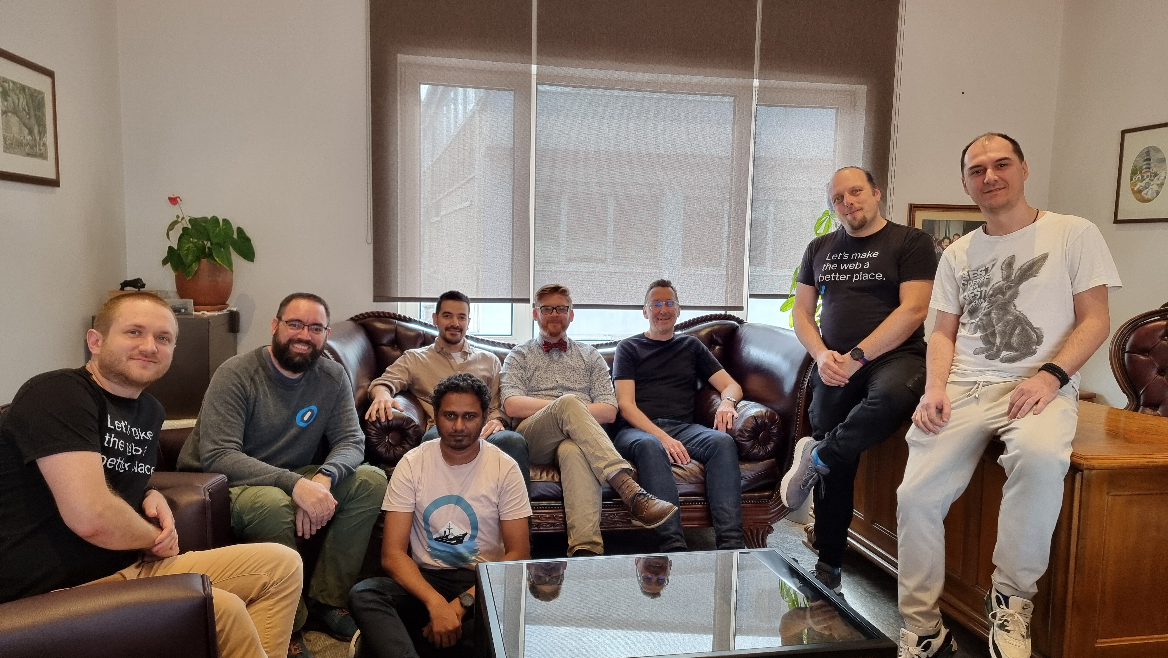 Full image of A group of men sit on chairs, a sofa, and the edge of a desk in a comfortable large office space.