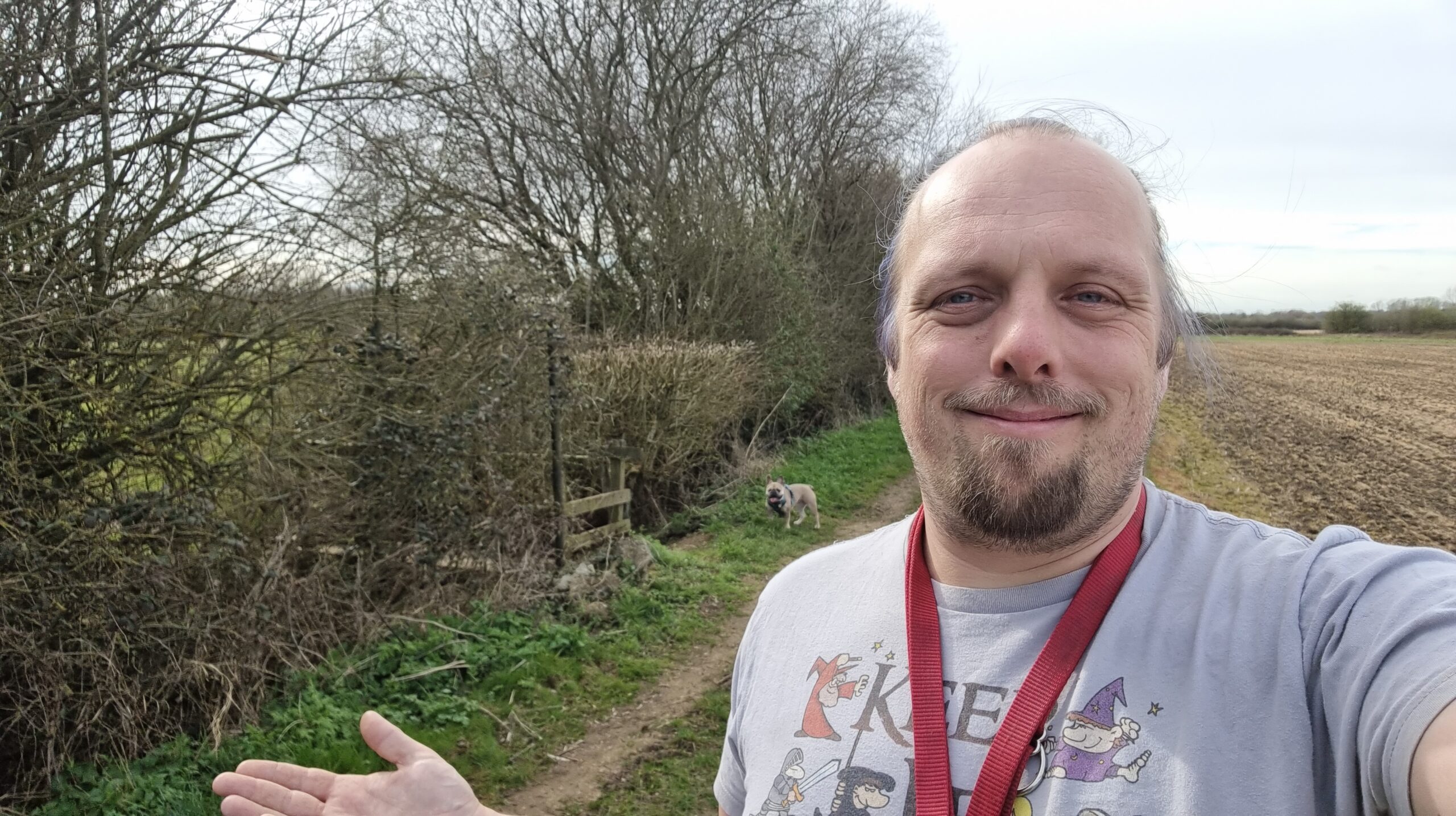 Full image of Dan, a red leash hung across his shoulders, gives a half-shrug as he smiles for a selfie on a grassy trail between a hedgerow and a field. Behind him, near a weathered old-fashioned footpath sign, his French Bulldog looks at him expectantly.
