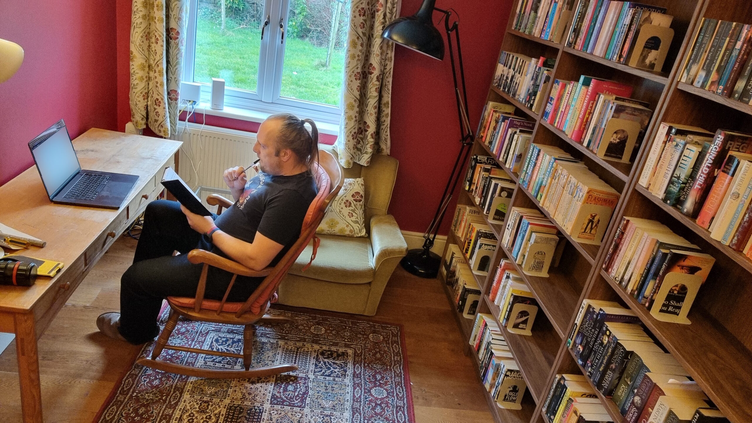 Full image of Dan, a white man with a ponytail, wearing a black shirt and jeans, sits in a rocking chair in front of an open laptop at a long desk; he has a notebook in his hand and holds a pen near his lips. He's in a domestic library with deep red walls, balanced-arm lamps, a woven rug on a wooden floor, and the wall behind him entirely covered with floor-to-ceiling bookshelves. A grassy lawn and sapling trees can be seen through a window, bordered by floral curtains and two clumsily-placed WiFi routers that sit on the window ledge. On the corner of the desk lie various hand tools, suggesting that light construction work has recently taken place.