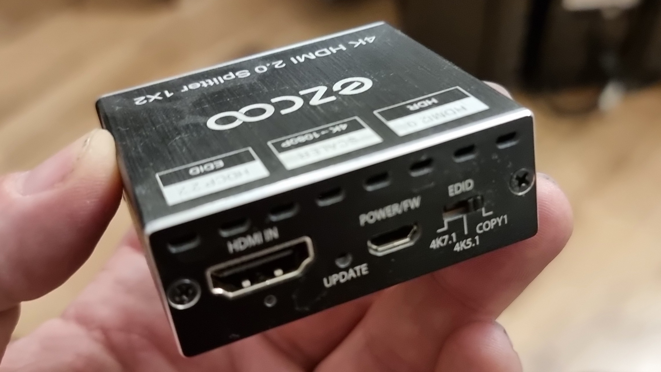 Full image of Fingers hold a small box, about half the size of a deck of cards, labelled "ezcoo 4K HDML 2.0 Splitter 1x2". On the side facing the camera can be seen a "HDMI In" port and an "EDID" switch that can be set to 4K7.1, 4K5.1, or COPY1.